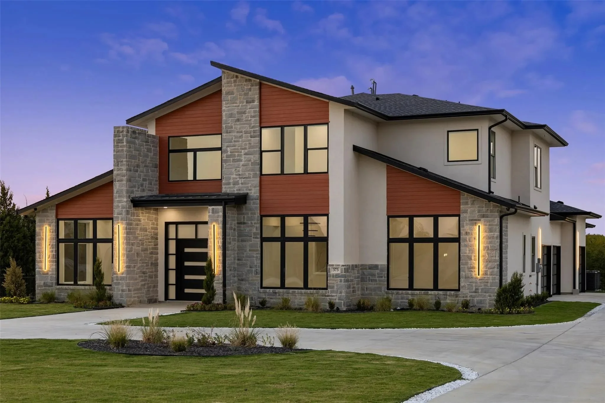 Modern two-story house with stone and wood exterior, large windows, and landscaped front yard at sunset.
