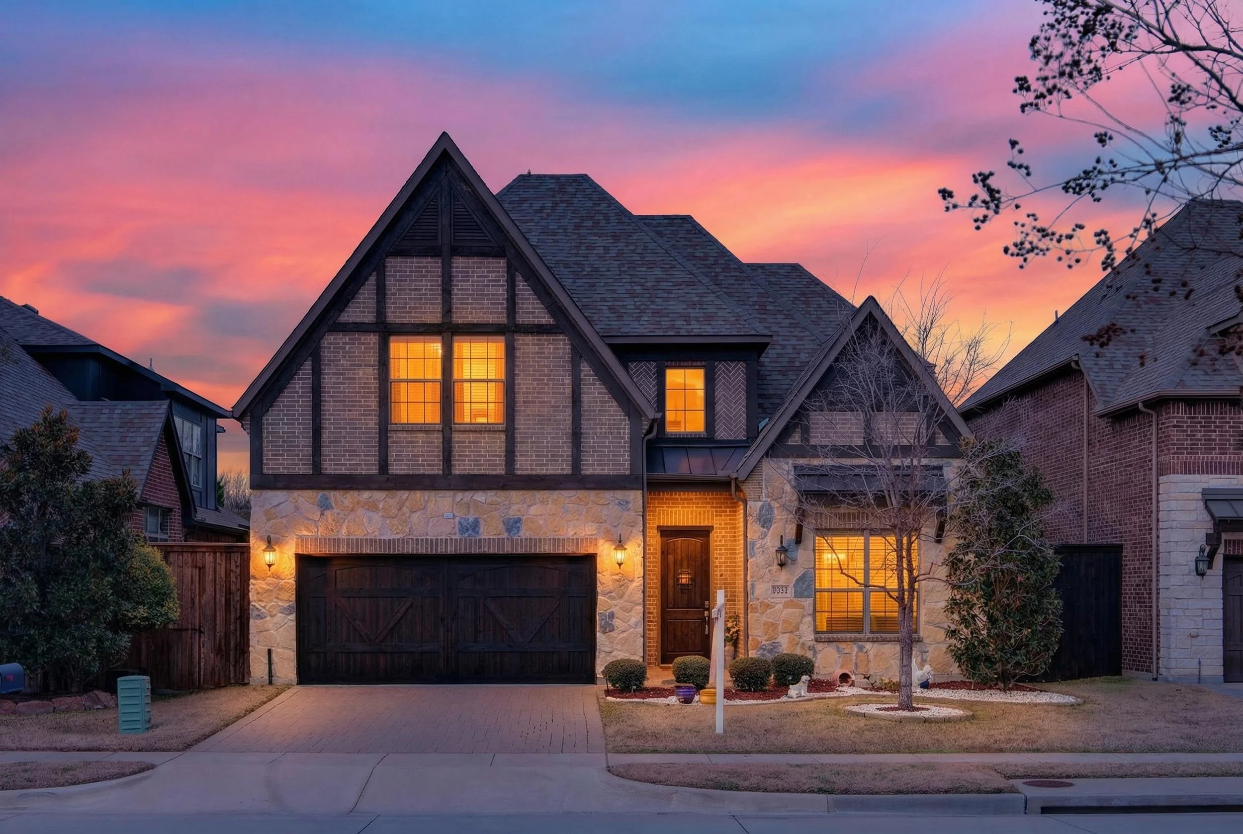 A two-story house with brick and stone exterior, illuminated windows, and a dark garage door, under a colorful sunset sky.