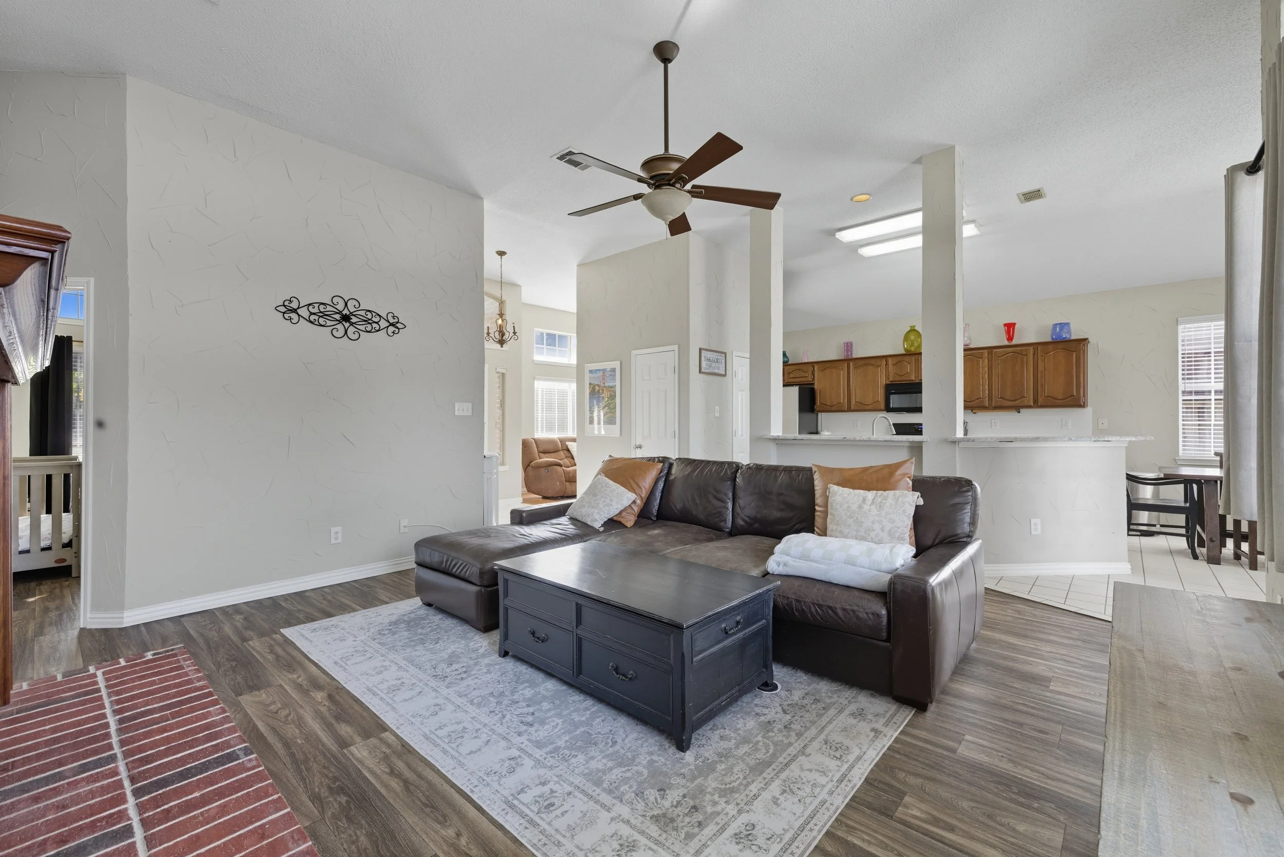 Living room with a dark brown leather sectional sofa, a black coffee table, a beige patterned rug, and a ceiling fan. The background includes a kitchen area with brown cabinets, a white countertop, and colorful vases on top.