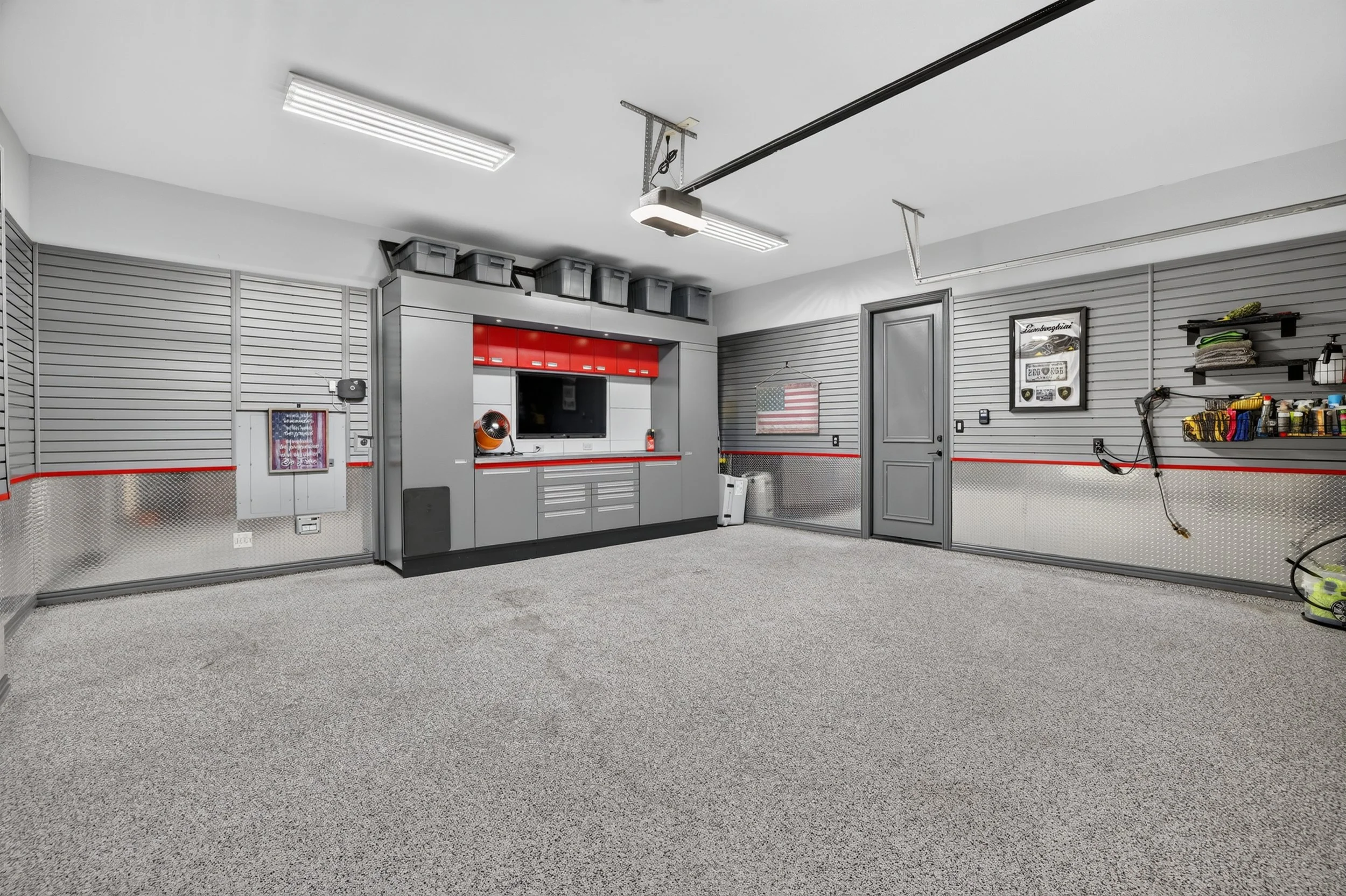 A clean, organized garage with gray slatwall panels, red and gray cabinets, a television, and various storage bins. The garage has fluorescent ceiling lights and a textured gray floor.