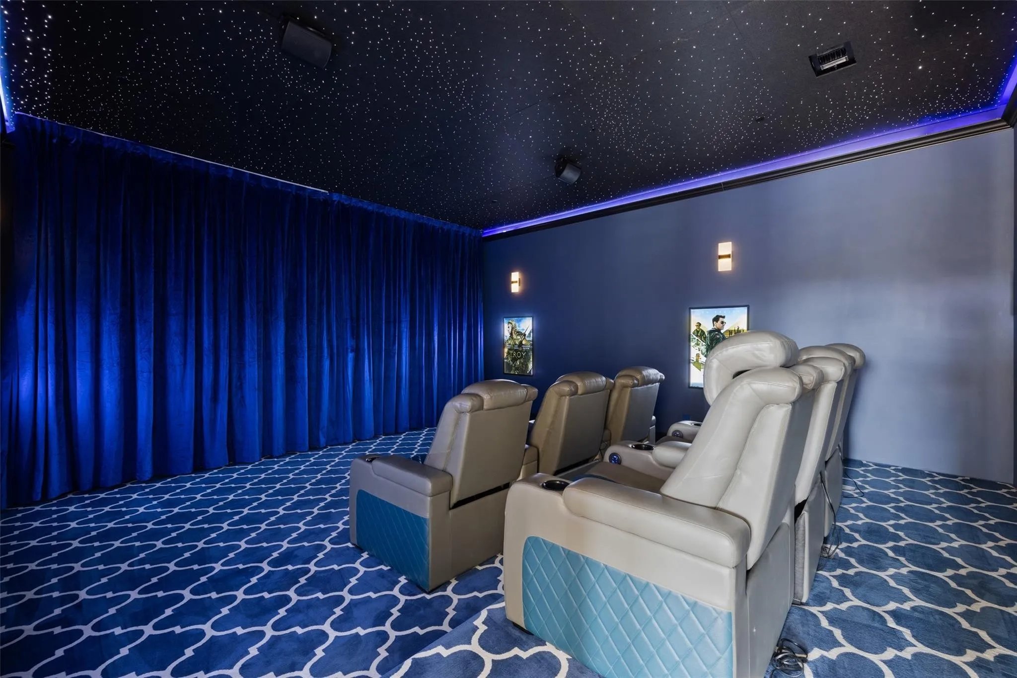 A private home theater with nine reclining chairs facing a wall with two screens, blue curtains, starry ceiling, and patterned carpet.