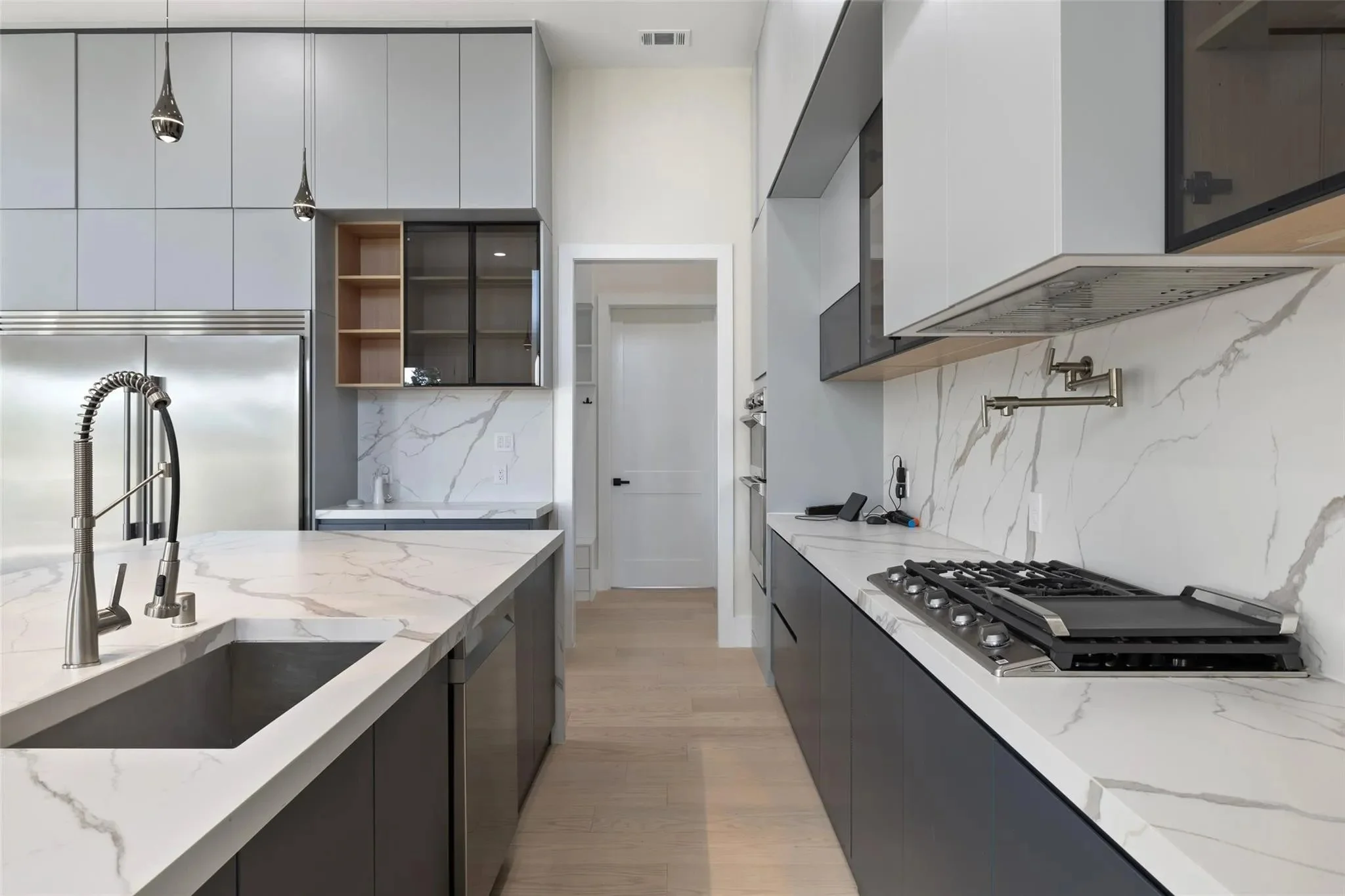 Modern kitchen with white marble countertops, black lower cabinets, and stainless steel appliances including a refrigerator and gas stove. There are pendant lights hanging from the ceiling and a small open shelving unit with glass doors.