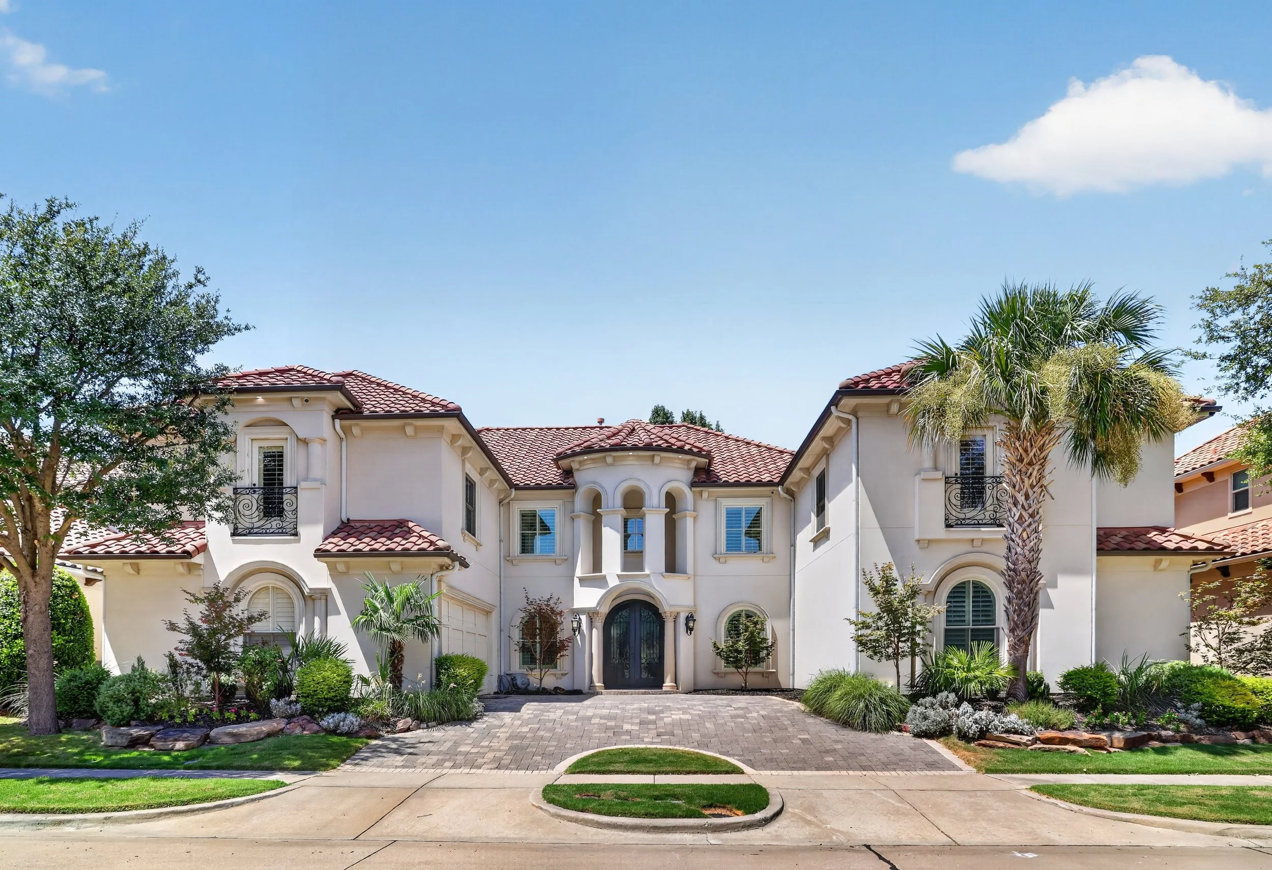 A large white house with red tile roof, front yard with landscaped bushes and trees, paved driveway, and a clear blue sky.