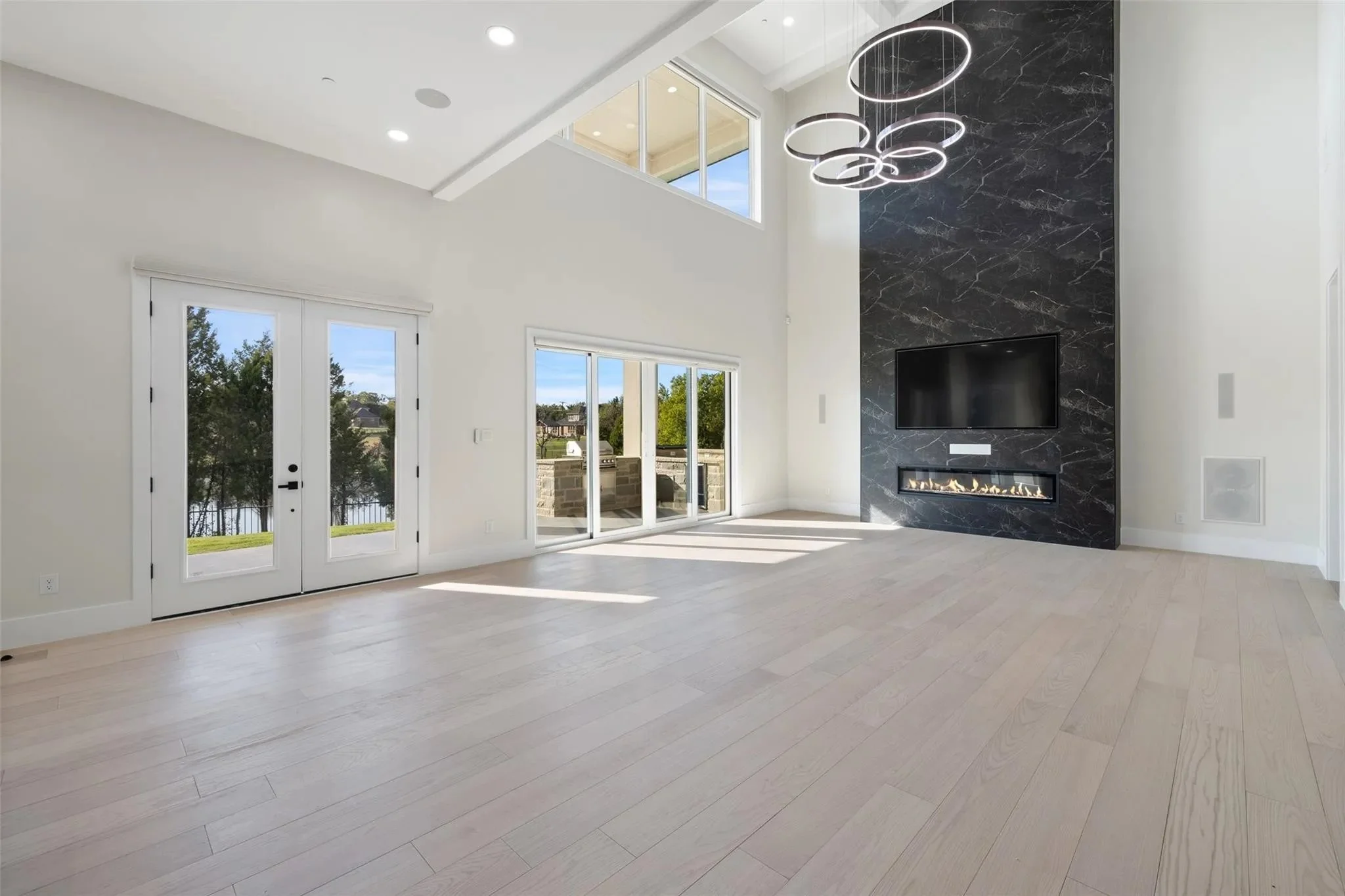 Empty modern living room with large windows, glass doors leading outside, a black marble accent wall with a mounted flat-screen TV, a linear fireplace, and a contemporary circular chandelier.