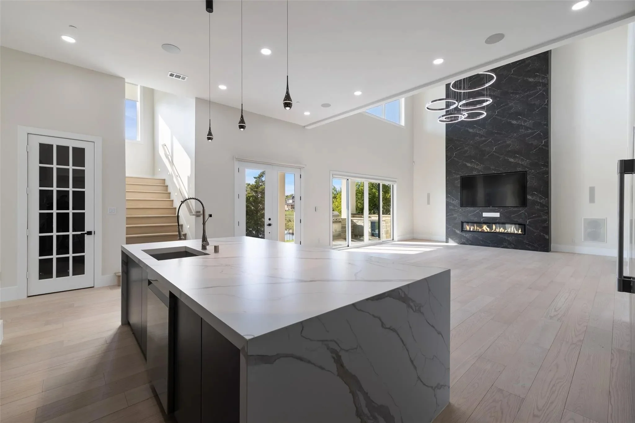 Modern open-concept living room with a kitchen island, black marble fireplace, and large windows showing outdoor scenery.