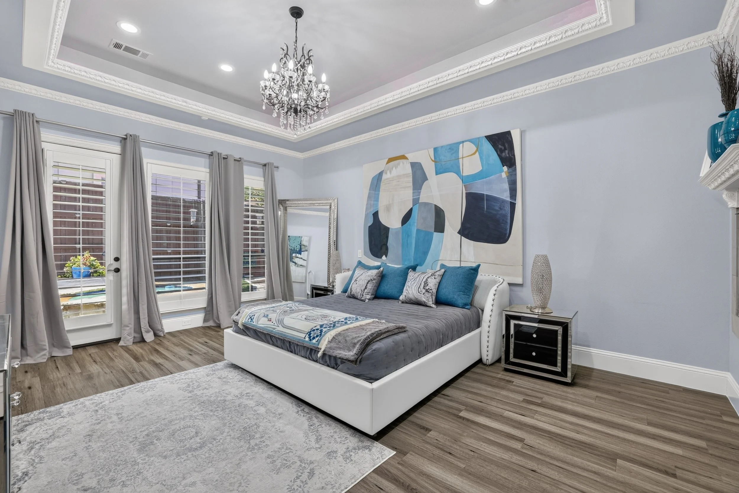 A bedroom with light blue walls, a white bed frame with gray and blue bedding, a large abstract blue and white painting above the bed, two black nightstands with table lamps, a chandelier hanging from the ceiling, gray curtains on a glass door, and wood flooring.