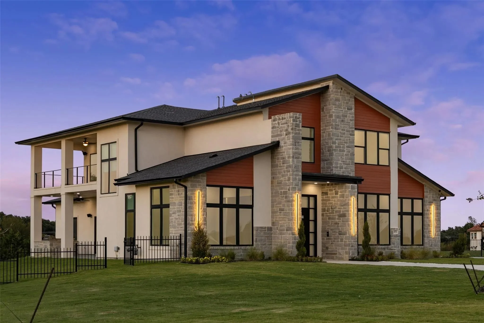Modern two-story house with large windows, stone accents, and a balcony at sunset.