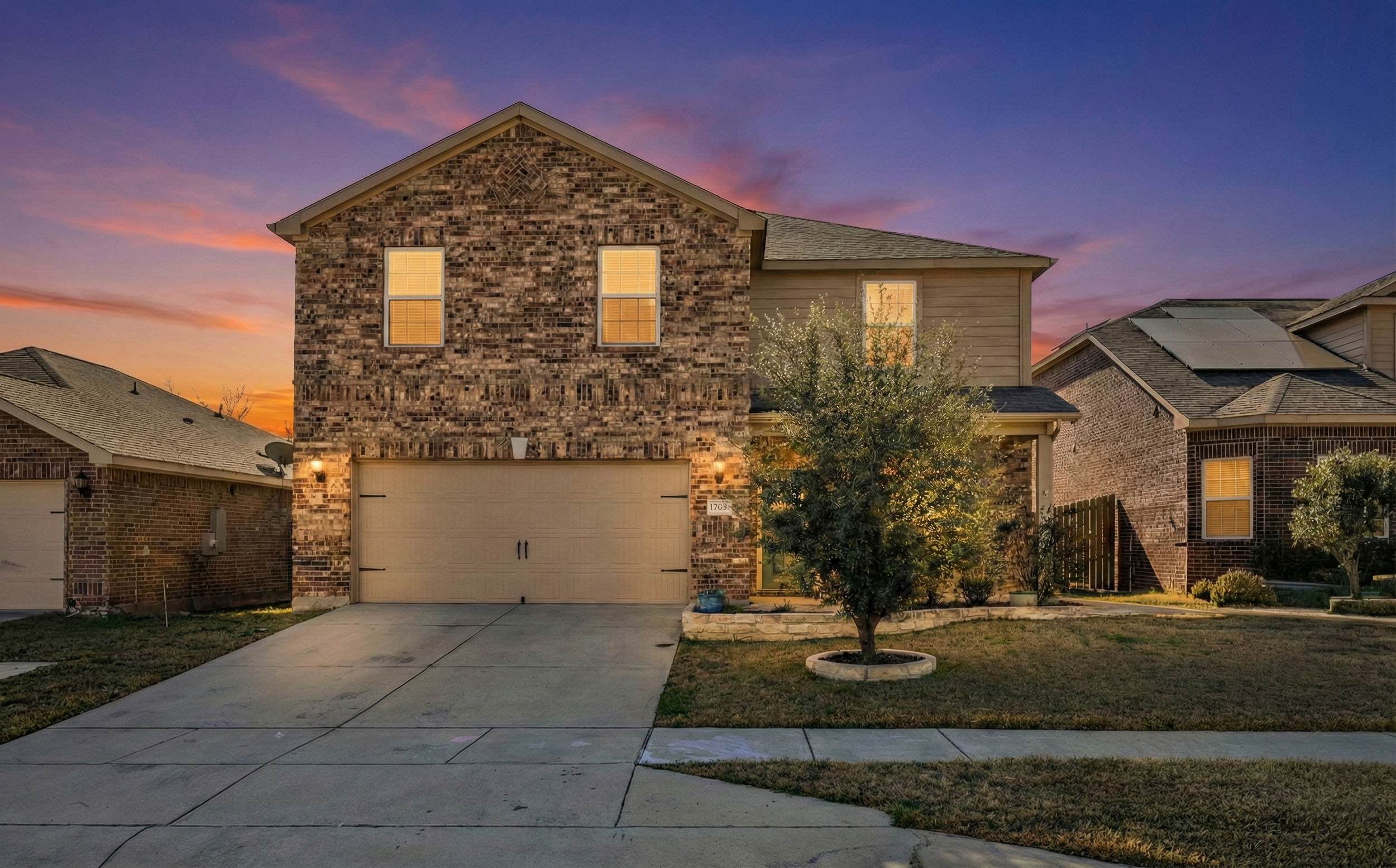 A suburban house at sunset with brick and beige siding, illuminated windows, a two-car garage, a small front yard with a tree and lawn, and neighboring houses