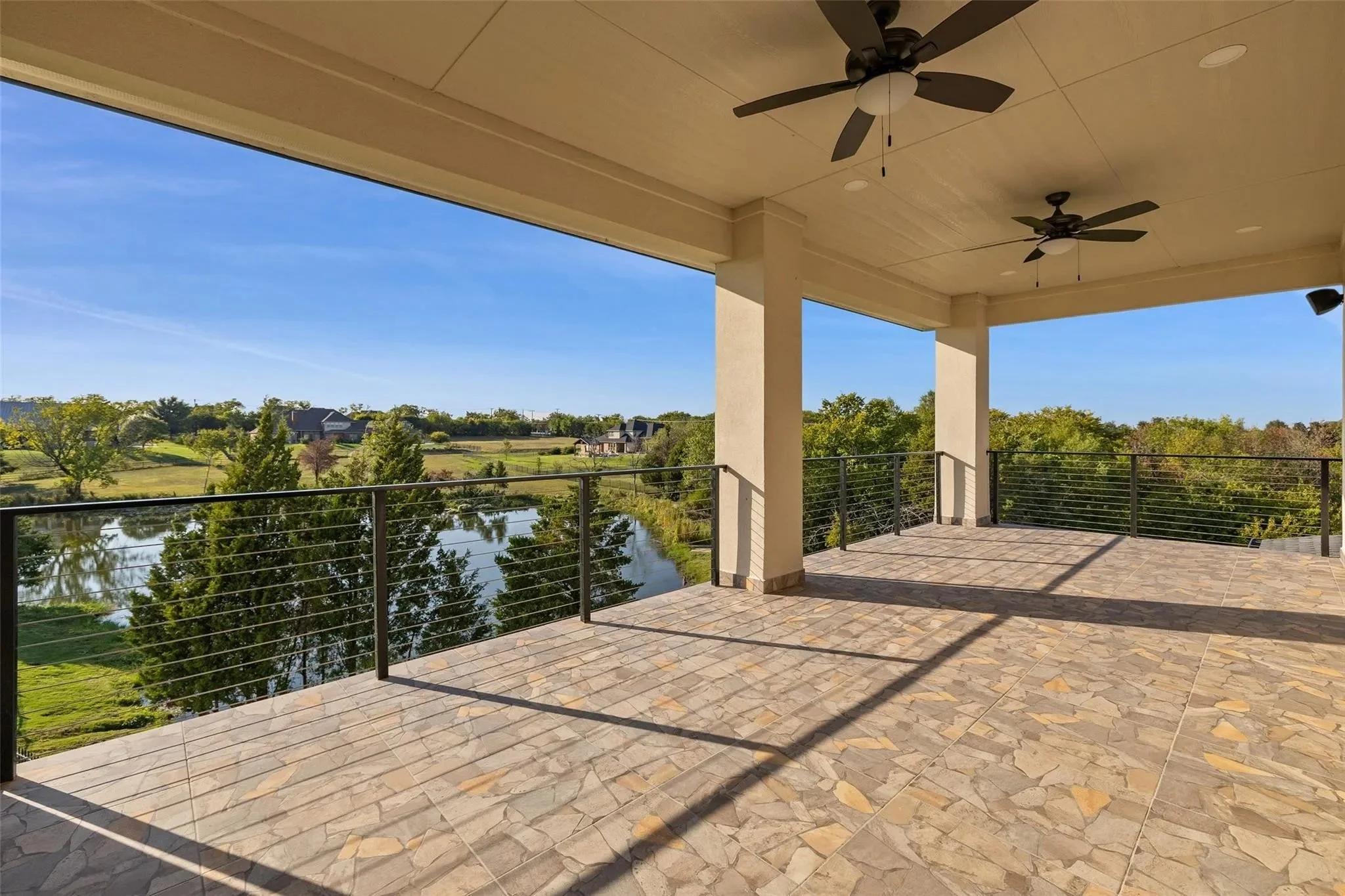 Empty outdoor patio with tile flooring, black metal railing, two ceiling fans, and a scenic view of trees, a pond, and distant houses under a clear blue sky.