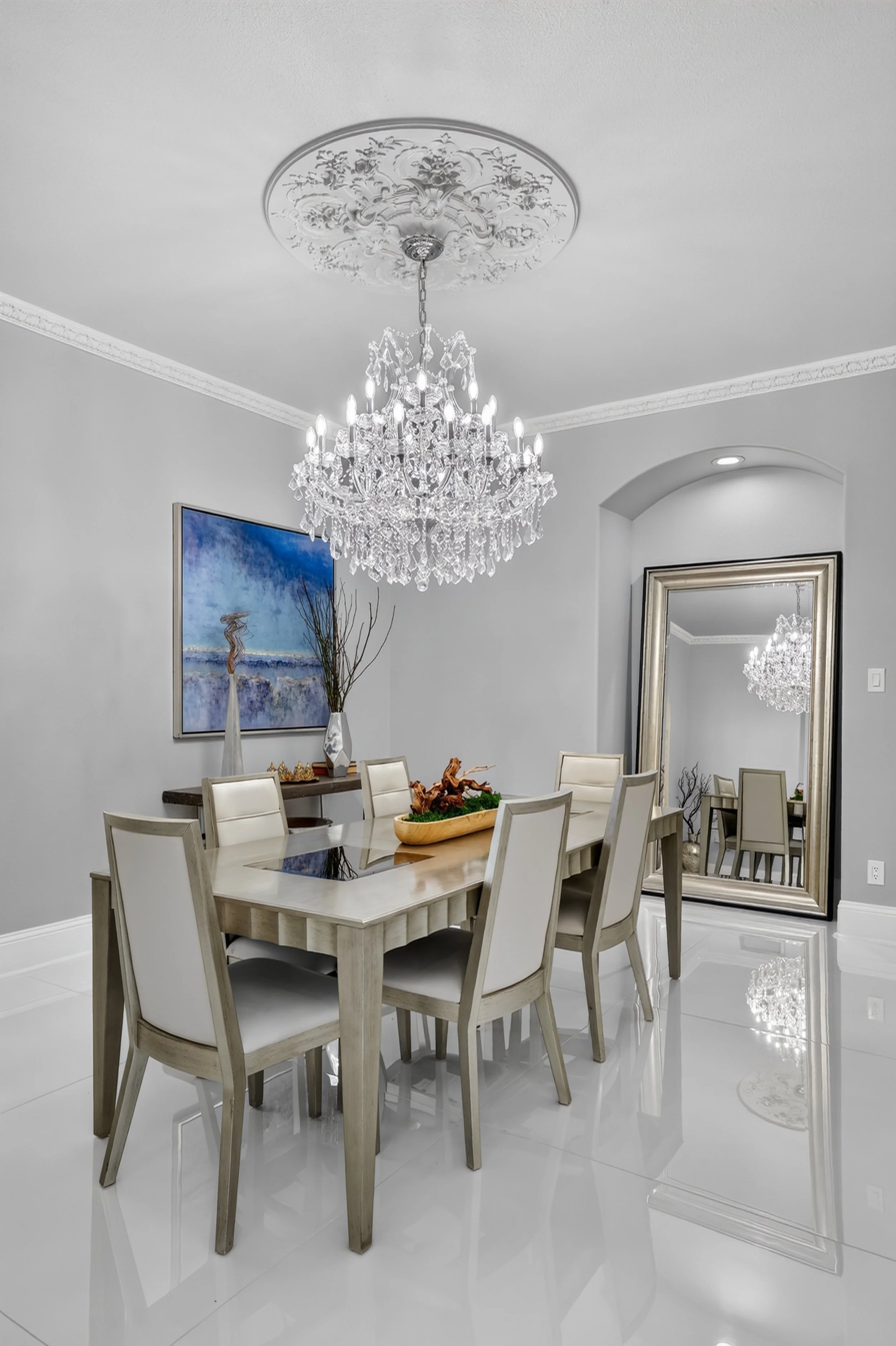 A dining room with a white table and cream-colored chairs, a large crystal chandelier hanging from an ornate ceiling medallion, a wall mirror, a painting, and decorative vases and sculptures.
