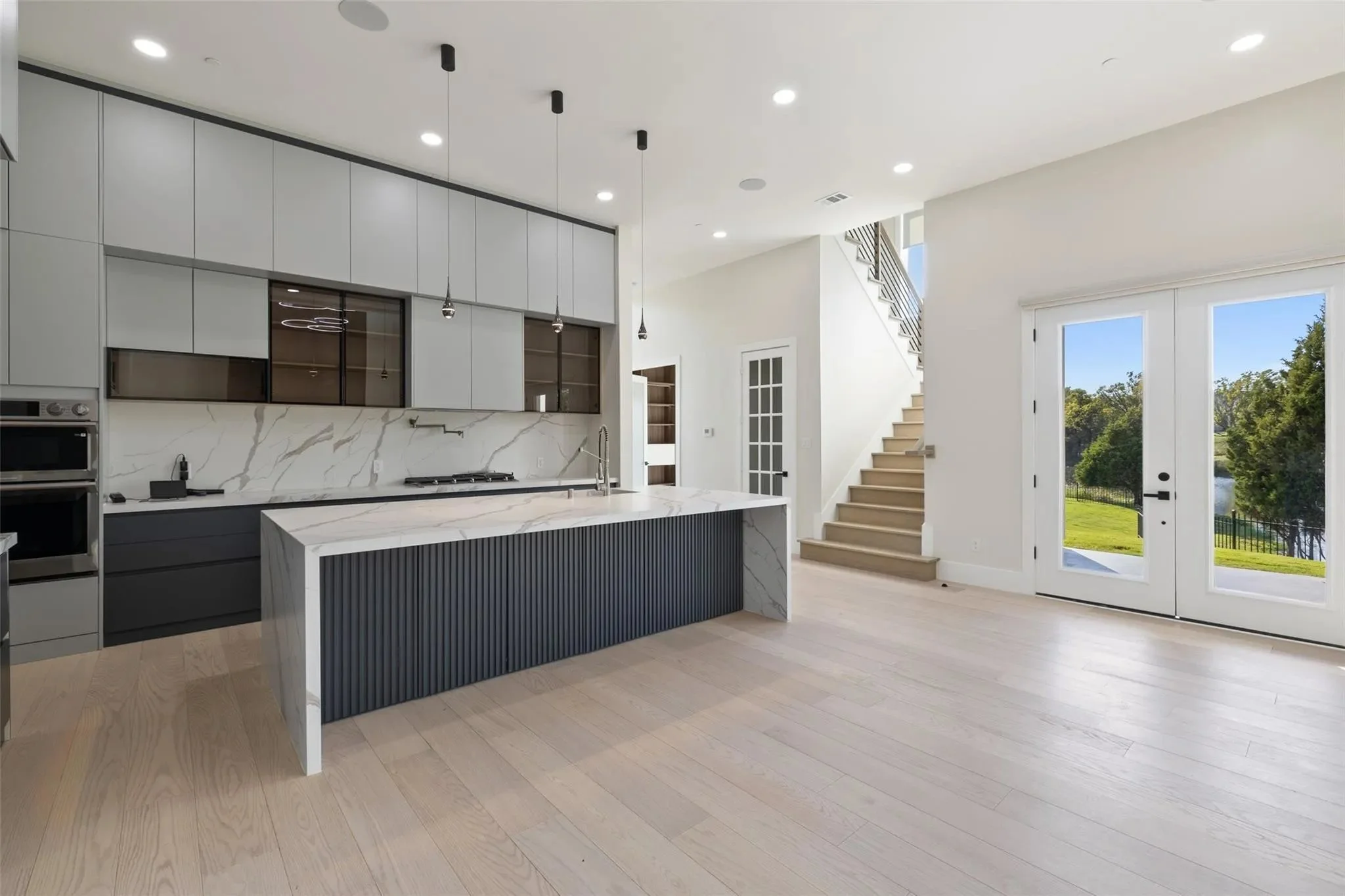 Modern kitchen with white and dark gray cabinetry, marble island with a patterned dark panel, light wooden flooring, large glass doors leading outside, and a staircase with light wooden steps and a black handrail.