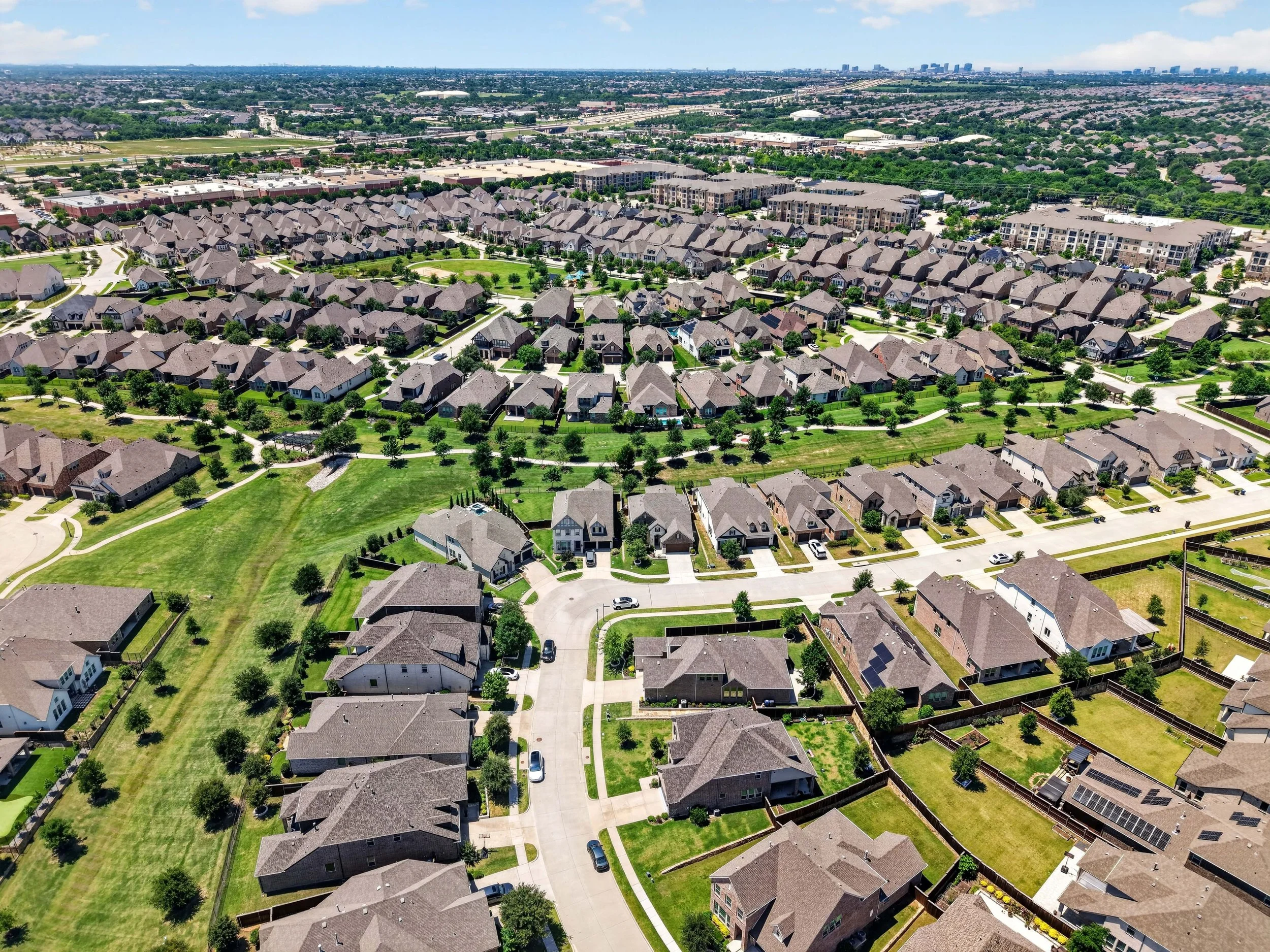 Aerial view of a suburban neighborhood with rows of houses, green lawns, and trees, with a golf course and city skyline in the background.