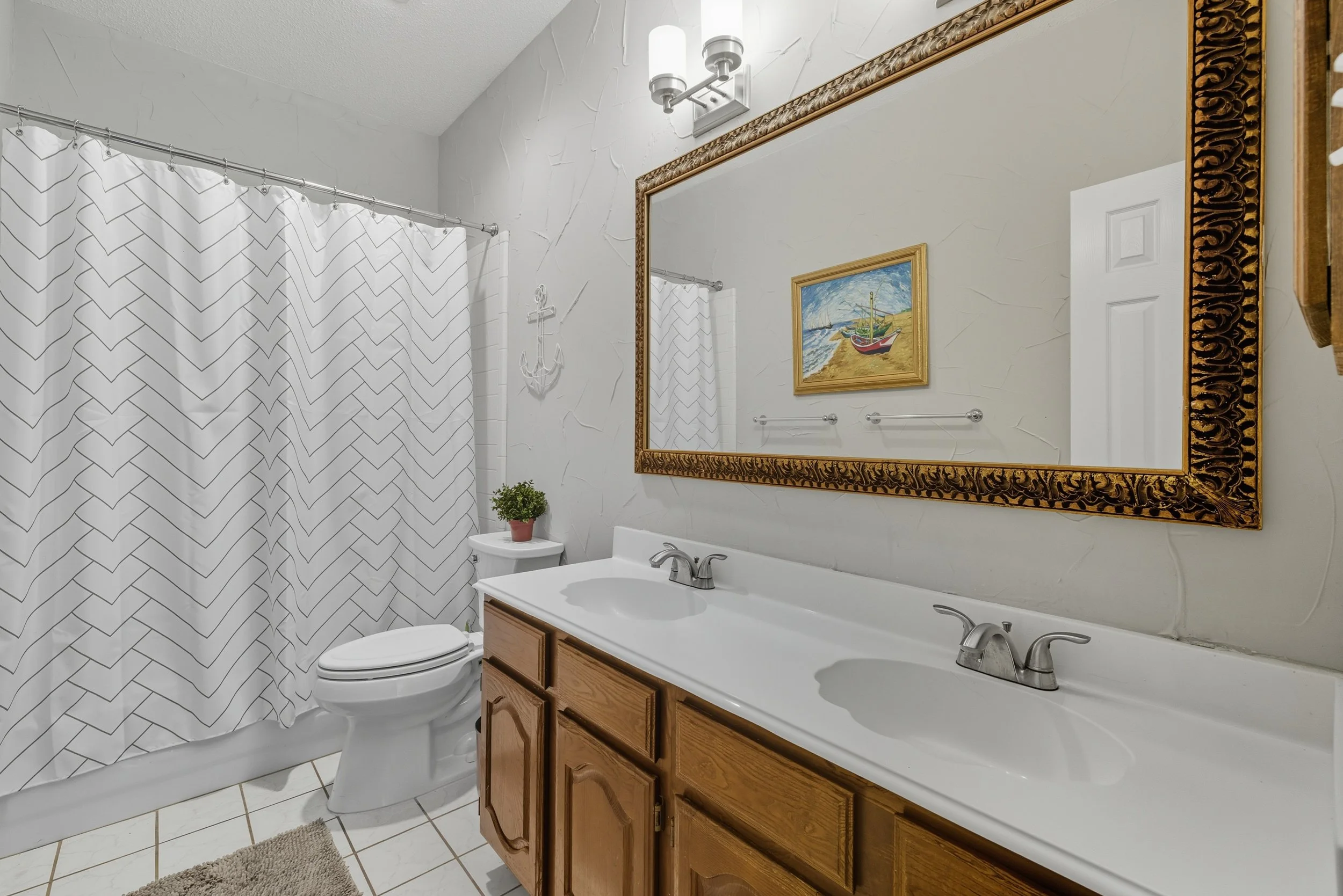 Bathroom with a double sink vanity, large mirror, white tile floor, potted plant, framed ocean scene painting, anchor wall decor, and a shower with chevron patterned curtain.