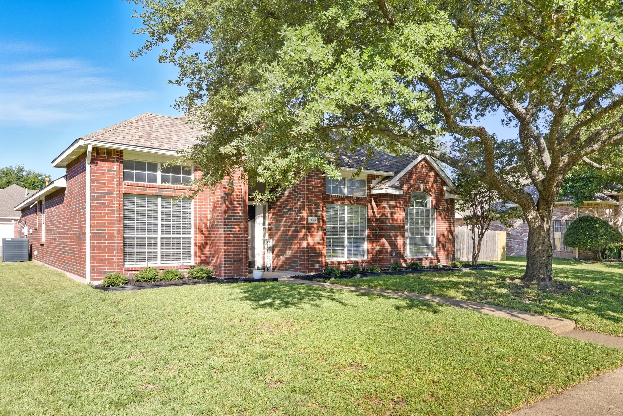 Red brick suburban house with large windows and a front yard shaded by a big tree under a clear blue sky.