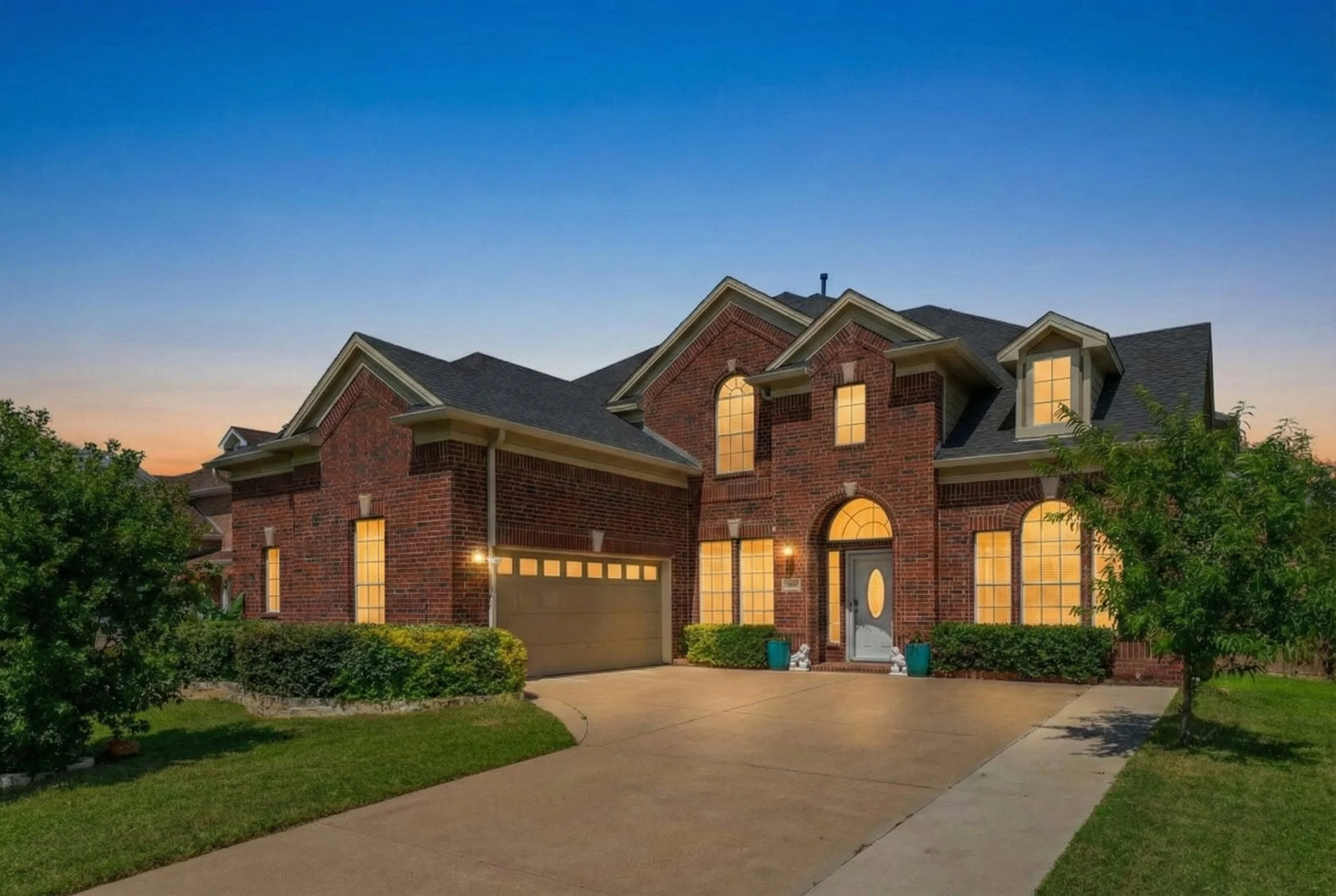 A large brick house with illuminated windows at dusk, surrounded by a well-maintained lawn and trees, with a driveway leading to a garage.