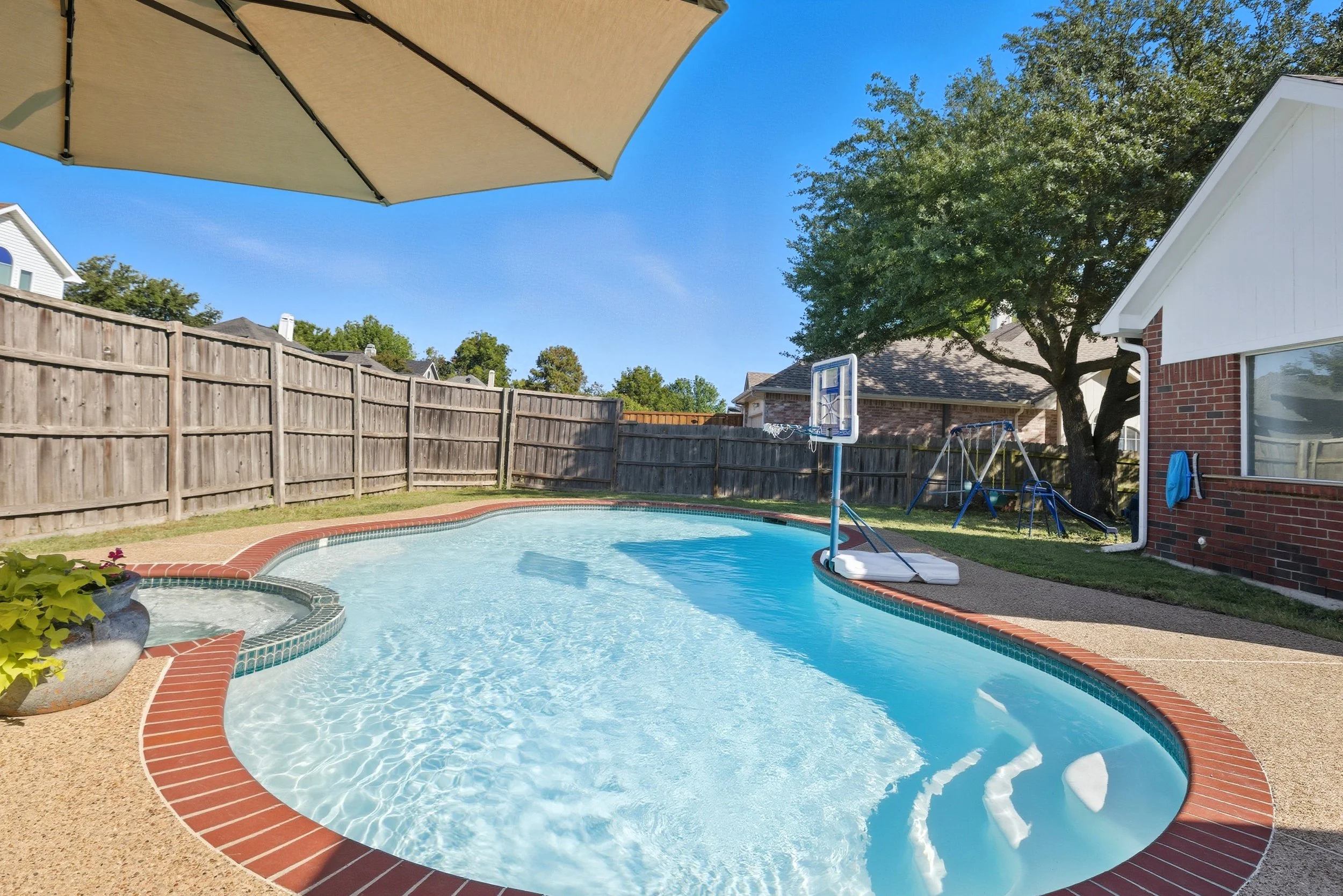Backyard with a swimming pool, an umbrella providing shade, a children's basketball hoop, and a swing set near a tree, with a wooden fence and neighboring houses in the background.