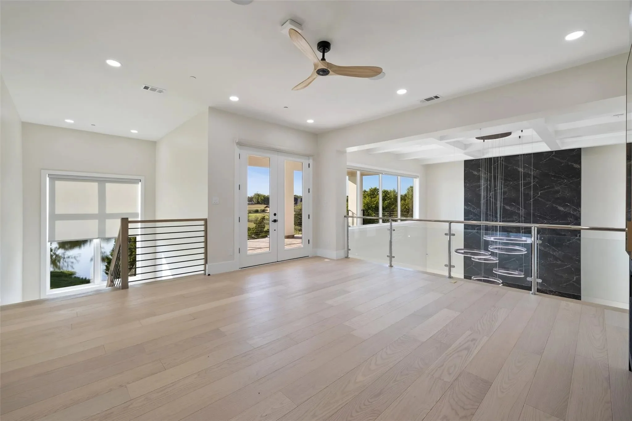 Spacious modern living area with light wood flooring, white walls, large windows, a glass door, a ceiling fan, and a black marble accent wall with a hanging circular light fixture.