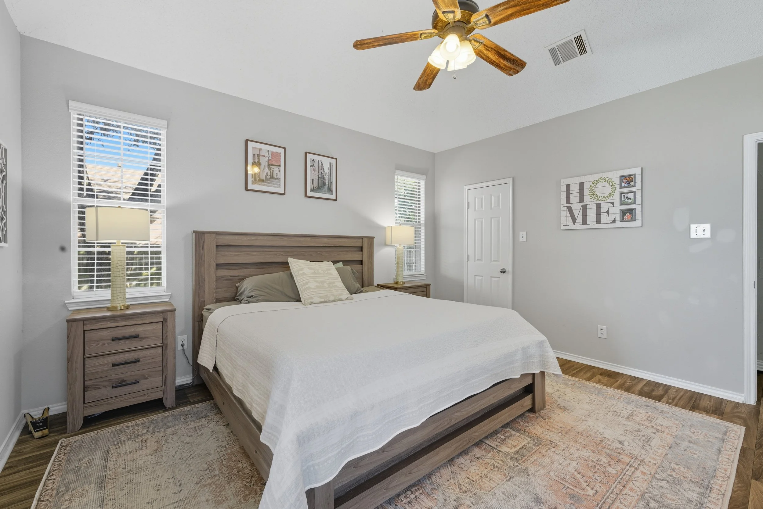 A bedroom with a bed, nightstands on each side with lamps, curtains on windows, wall art, a ceiling fan, and a door.
