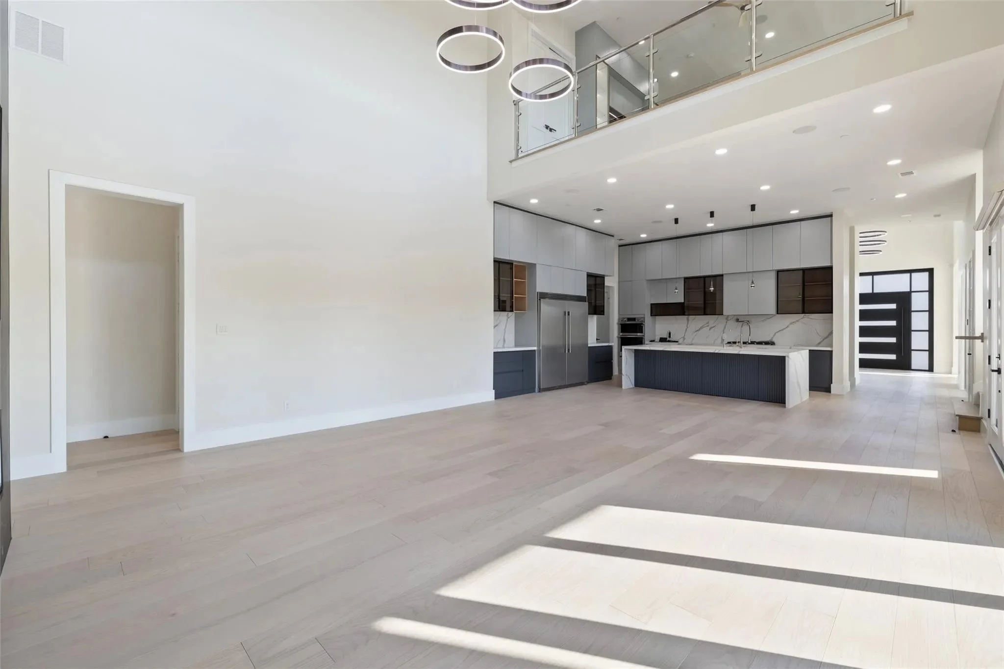 Spacious, modern open floor plan kitchen and living area with light wood flooring, white walls, and large windows letting in natural light. The kitchen features a large island, gray cabinets, and stainless steel appliances. Modern lighting fixtures h