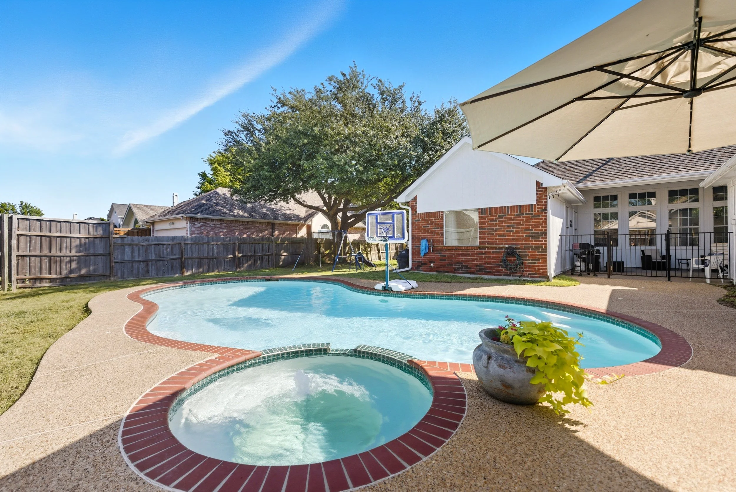 Backyard with swimming pool, hot tub, patio, large tree, house with brick wall, outdoor furniture under a large umbrella, and a clear blue sky.