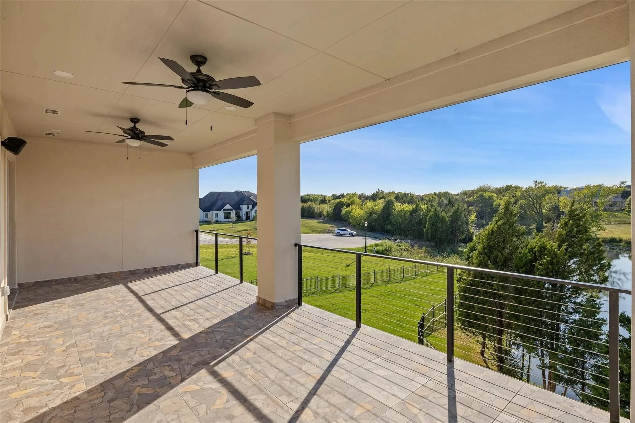 Balcony with tiled floor, ceiling fans, black railing, overlooking green lawn, trees, small pond, and houses in the distance under a clear blue sky.