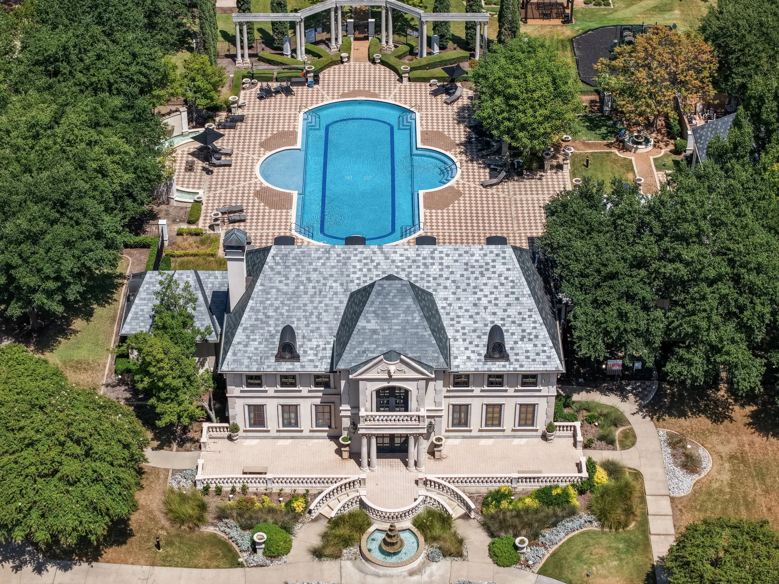 An aerial view of a large mansion with a swimming pool in the backyard, surrounded by lush green trees and landscaping.