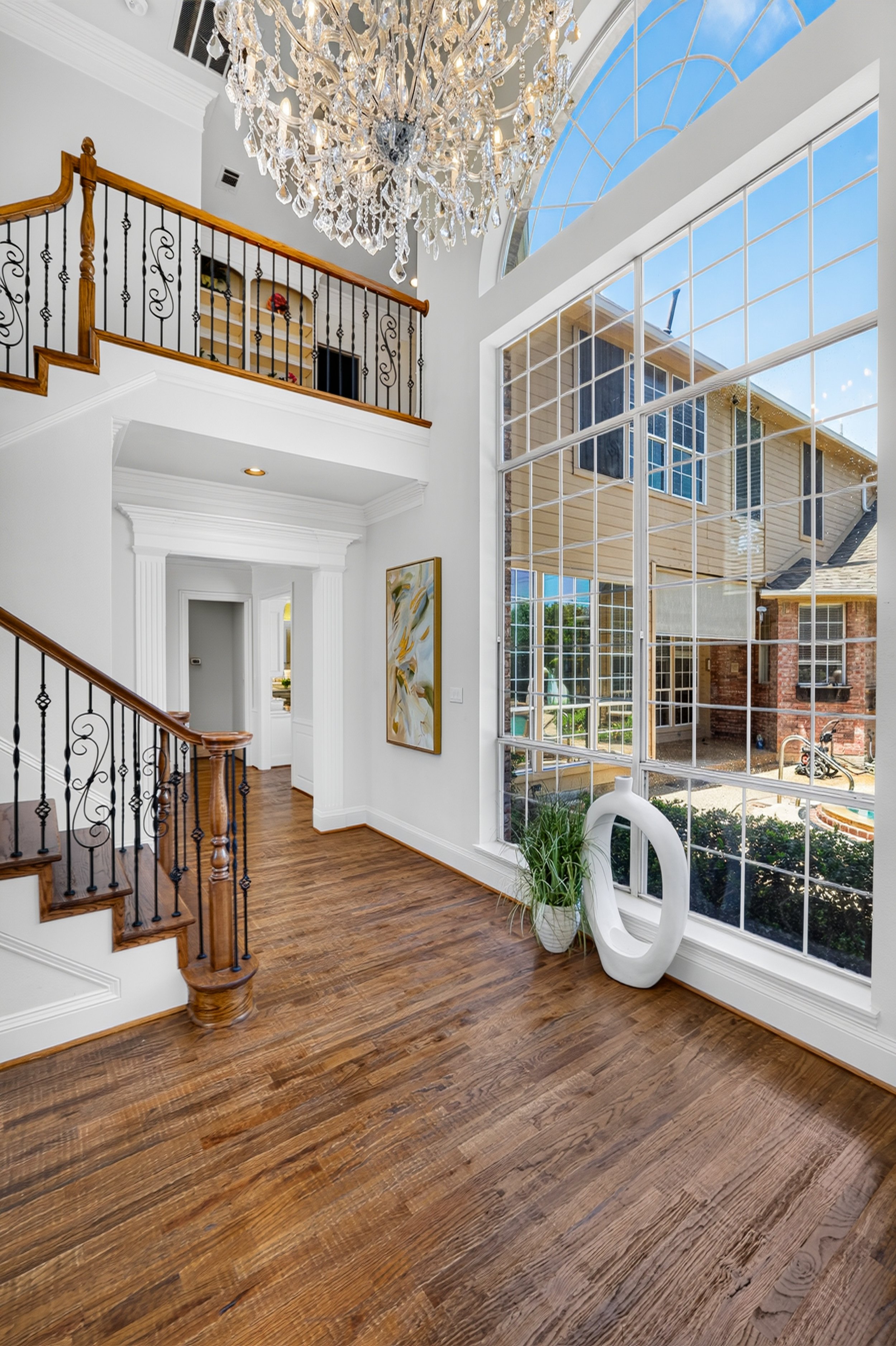 Interior view of a spacious living room with a large window, a crystal chandelier, hardwood floors, and a staircase with wooden handrails and decorative black iron balusters.