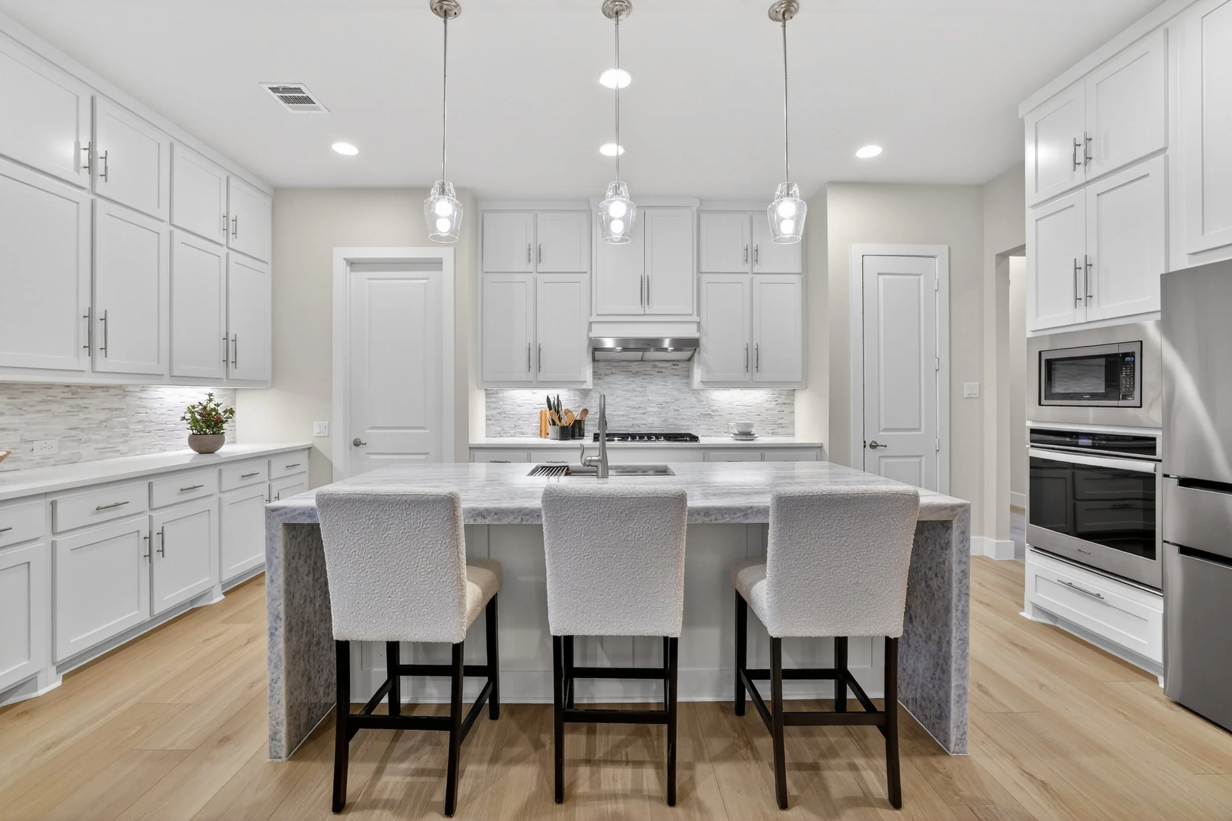 Modern kitchen with white cabinets, marble island with four upholstered chairs, stainless steel appliances, and pendant lighting.