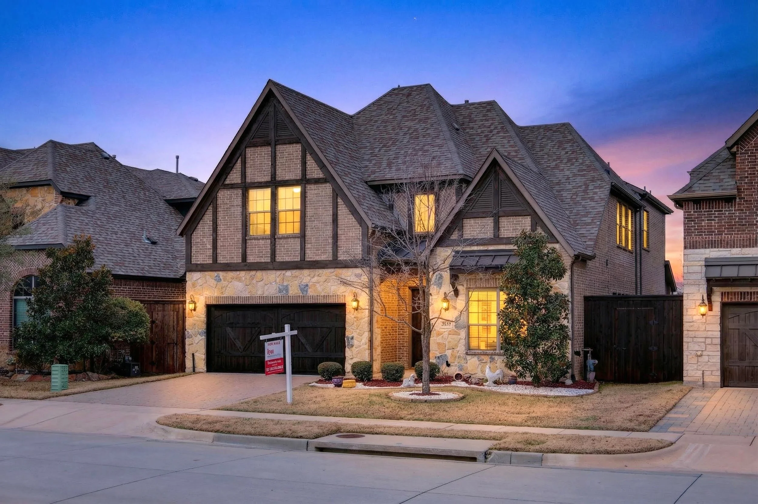 A large, multi-story house with brick and stone exterior, illuminated windows, and a decorative front yard with trees and a 'For Sale' sign.