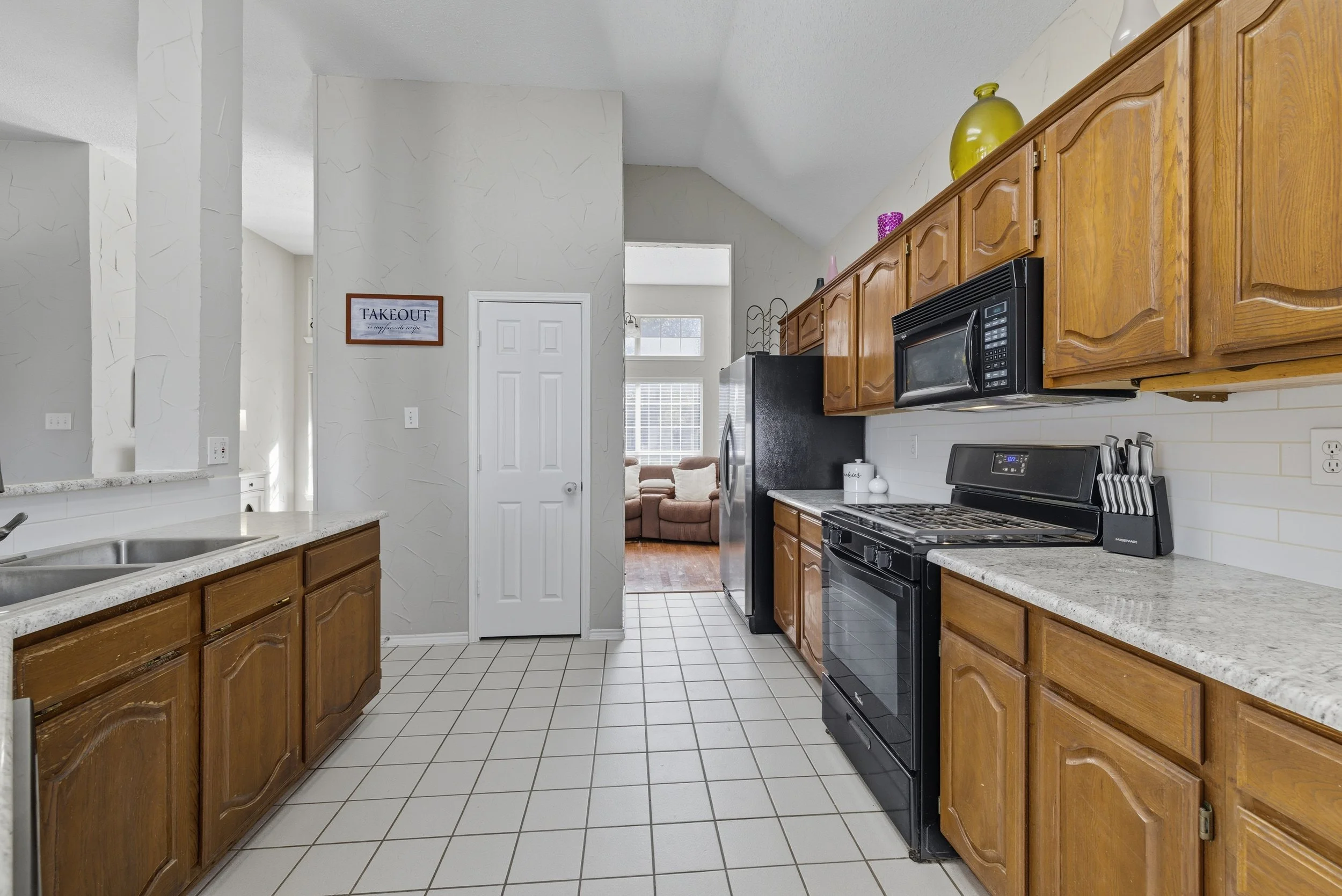 Kitchen with wooden cabinets, black appliances, granite countertops, white tiled floor, and a view into a living room with a white sofa and windows.