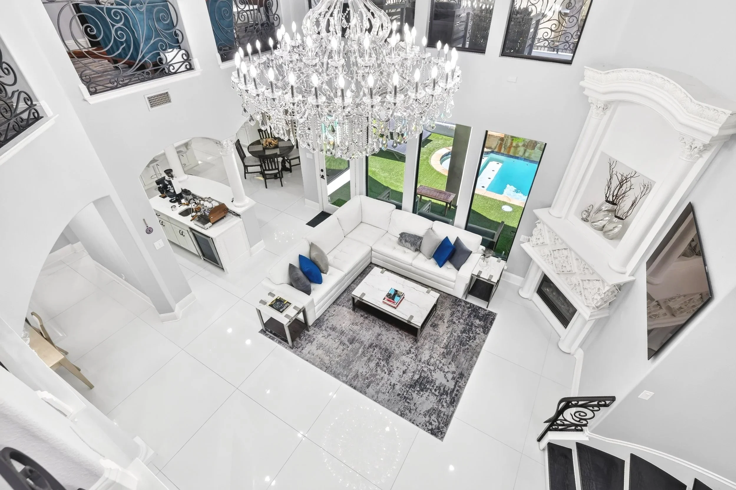 Living room with a large crystal chandelier, white sectional sofa with colored pillows, glass sliding doors to outdoor pool area, decorative mantel with silver vases, white tiled floors, high ceilings, and large windows with iron balconies.