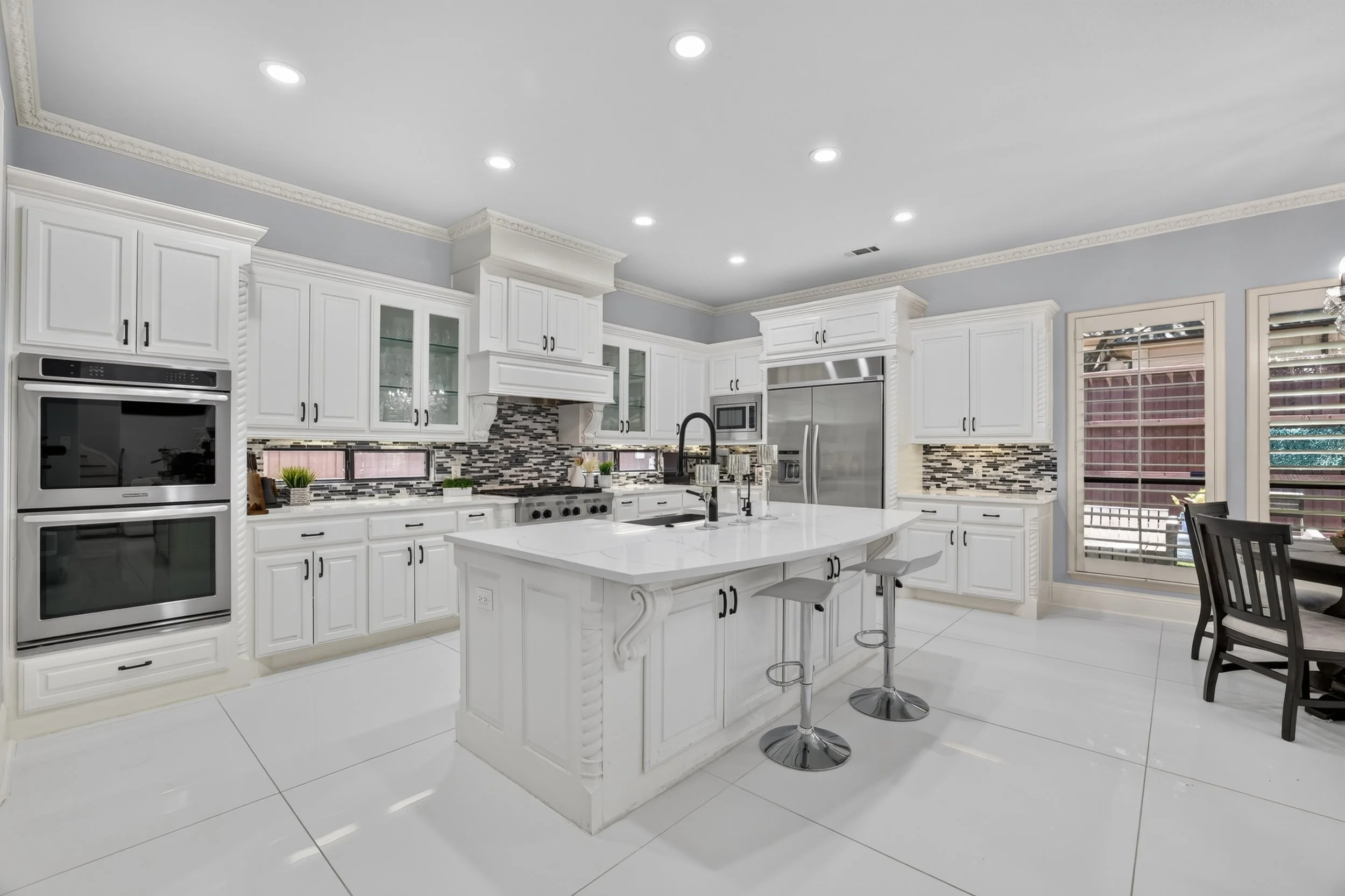 Modern white kitchen with an island, black hardware, mosaic tile backsplash, stainless steel appliances, crown molding, and large windows.