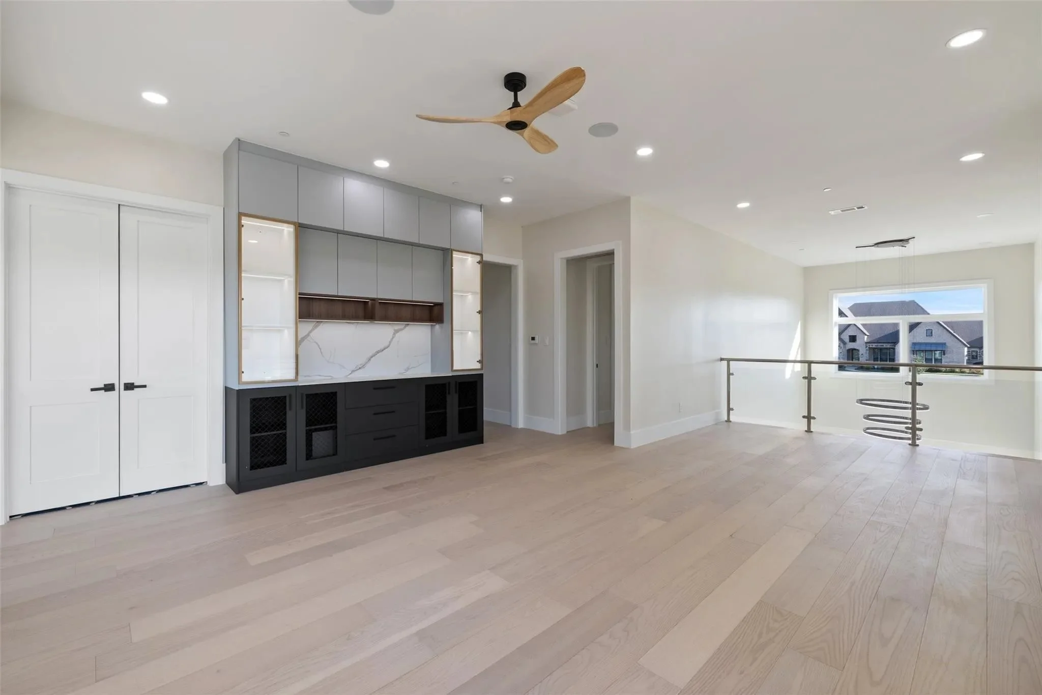 Empty living room with built-in shelves, large window view of neighboring houses, light-colored hardwood floors, white walls, and a modern ceiling fan.