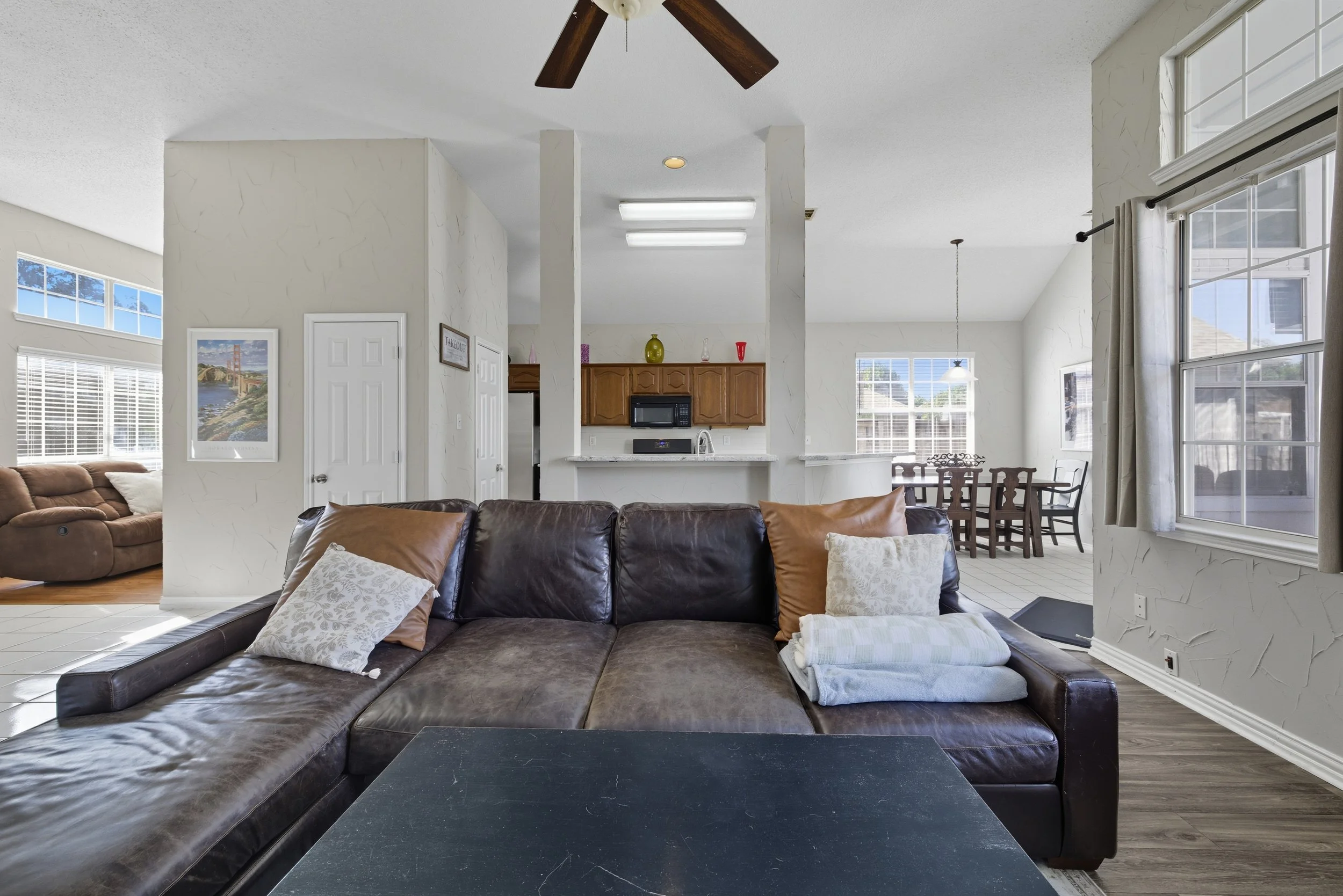 Living room with brown leather sofa, beige pillows, and a shelf with lamps and artwork visible in a well-lit residential space.
