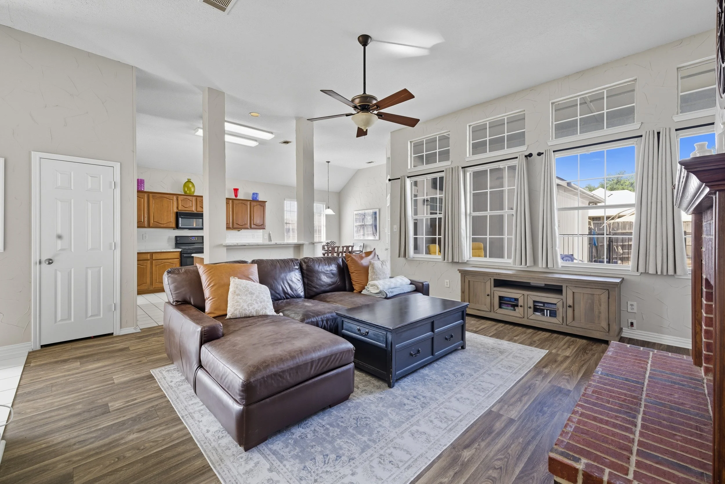 Living room with a brown leather sectional sofa, a black coffee table, and a wooden media console beneath large windows with white curtains. There is a ceiling fan and a brick fireplace in the room. The adjacent kitchen has wooden cabinets and a dini