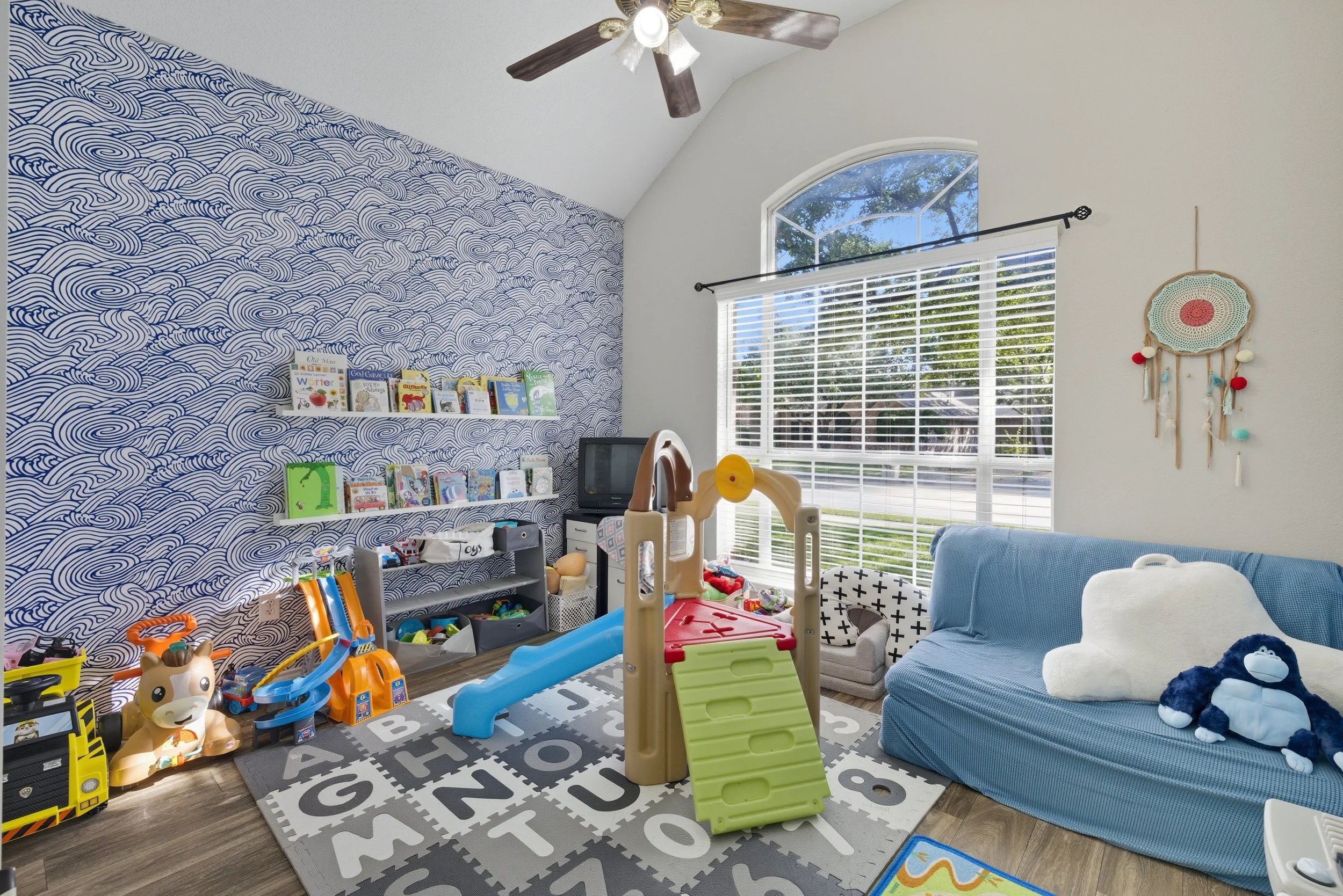 A children's playroom with blue and white decor, featuring a blue sofa with stuffed animals, a small slide and play structure, a toy storage unit, and a patterned rug with alphabet letters. There are books on the wall, a small TV, and a decorative wa