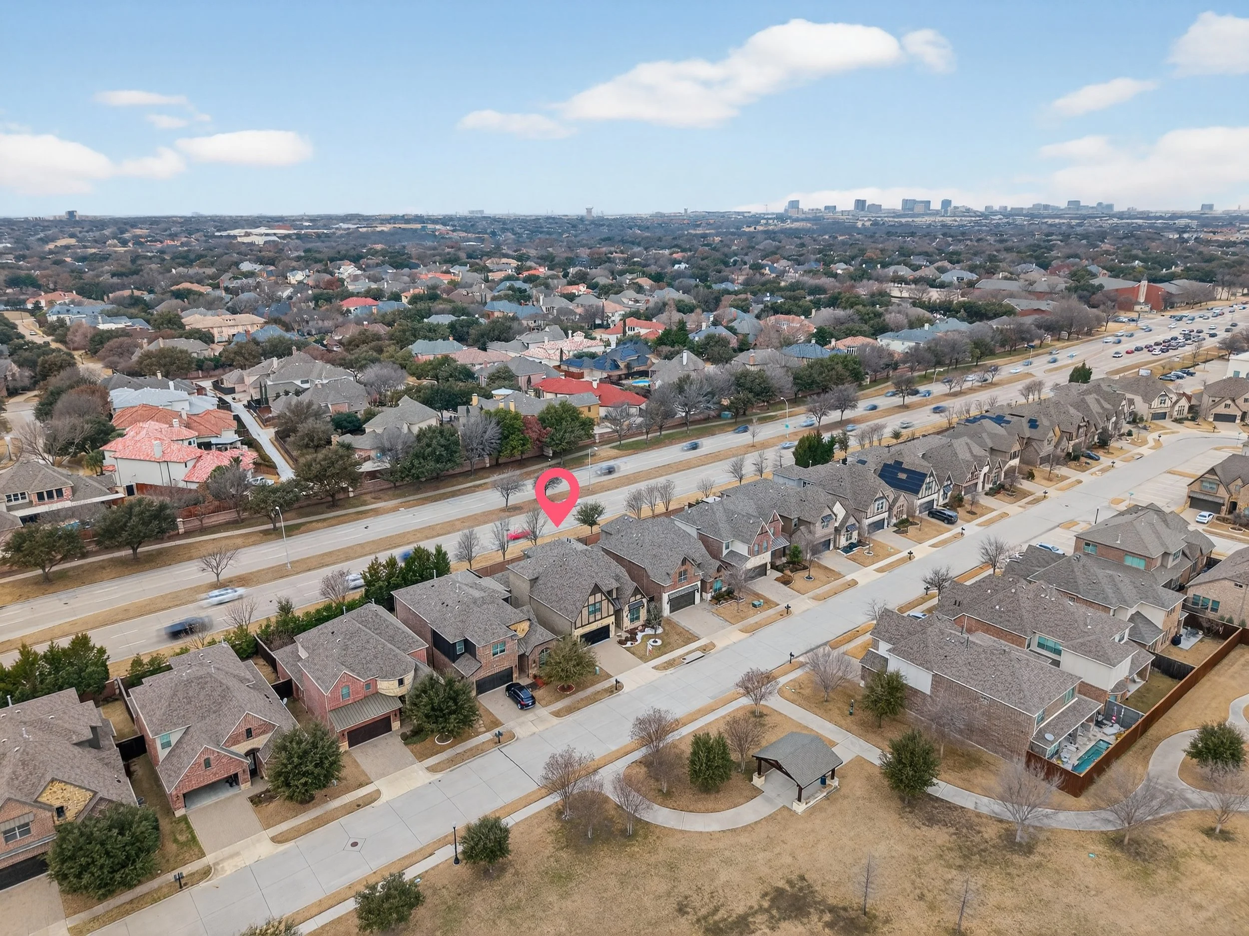 Aerial view of a suburban neighborhood with rows of houses, some with solar panels, leafless trees, and a main road with traffic. A pink location marker is over a specific house.