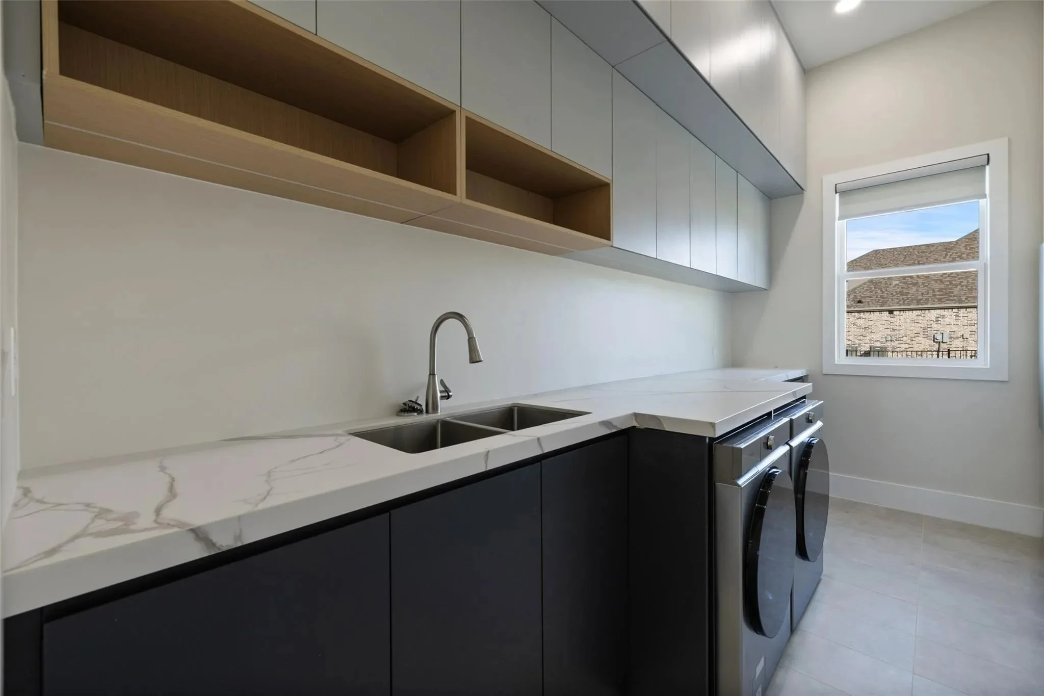 Modern laundry room with white marble countertop, black cabinetry, stainless steel sink, and wooden open shelves, with a window showing rooftops outside.