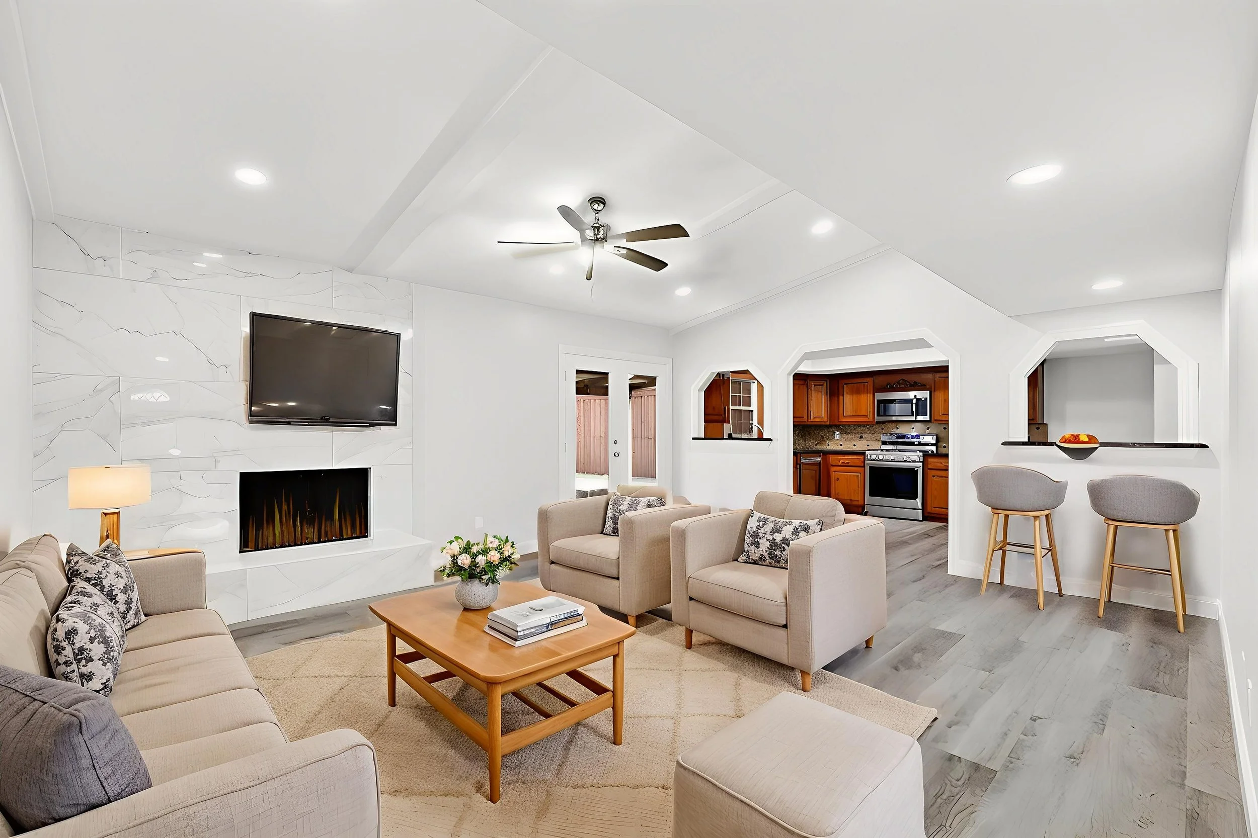Bright living room with white walls, beige sofas, a wooden coffee table with a flower arrangement, a TV mounted above a modern fireplace, and an open kitchen with wooden cabinetry.