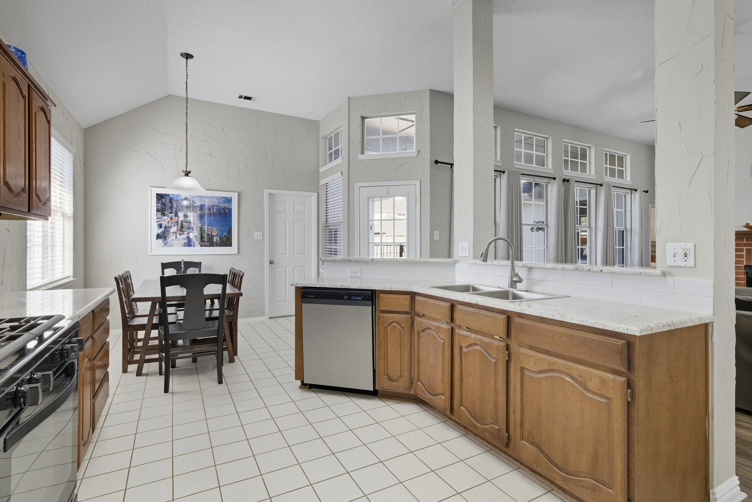 Kitchen with wooden cabinets, white countertops, and a dining area with a table and chairs, windows with curtains, and a painted wall with a landscape picture.