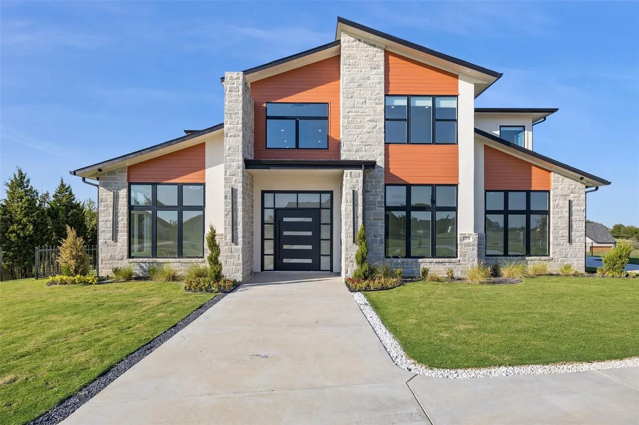 Modern two-story house with large windows, a concrete driveway, landscaped front yard, and a blue sky.