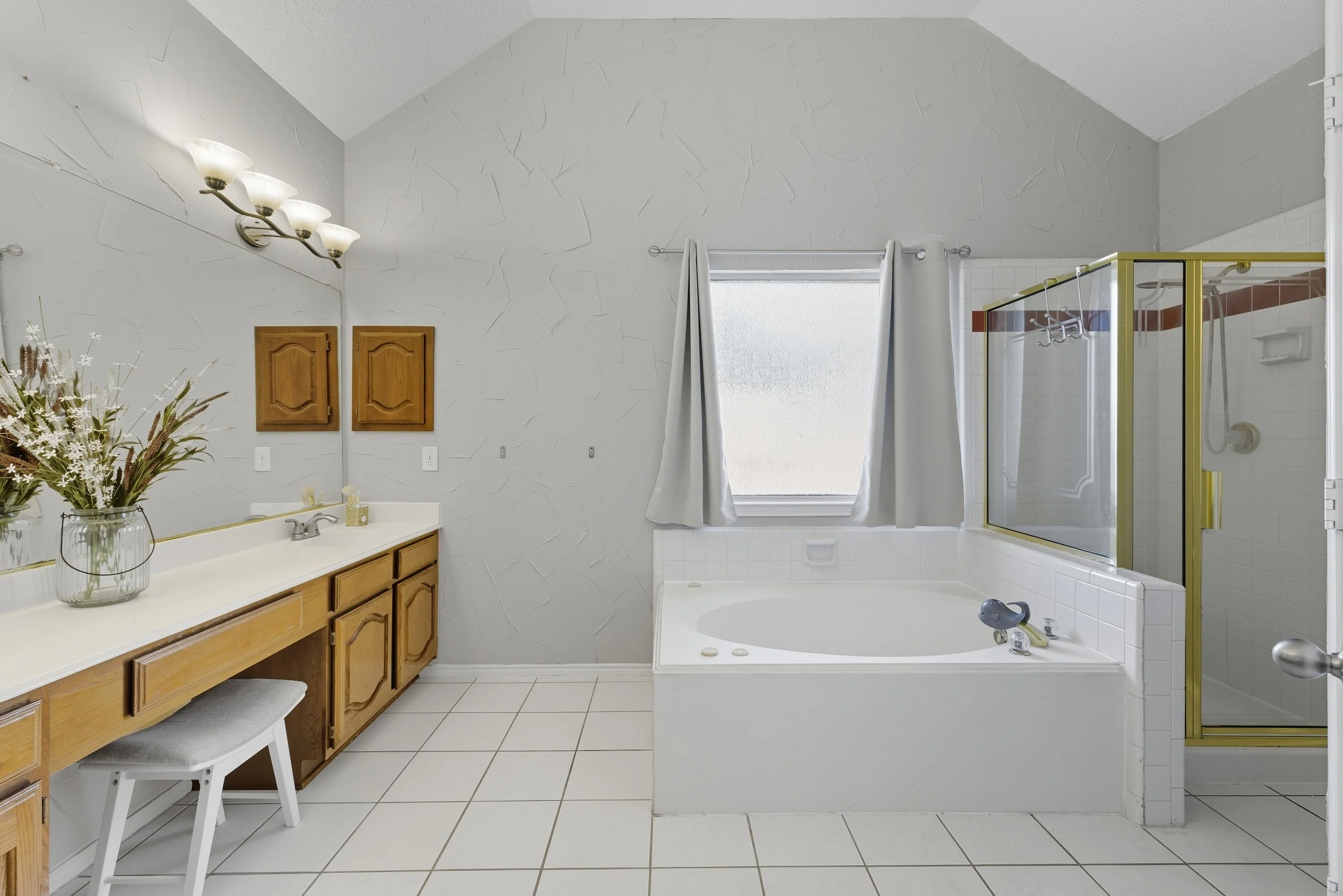 Bathroom with a large mirror, a wooden vanity with a sink, a bathtub with a window above, and a glass shower enclosure with gold trim.
