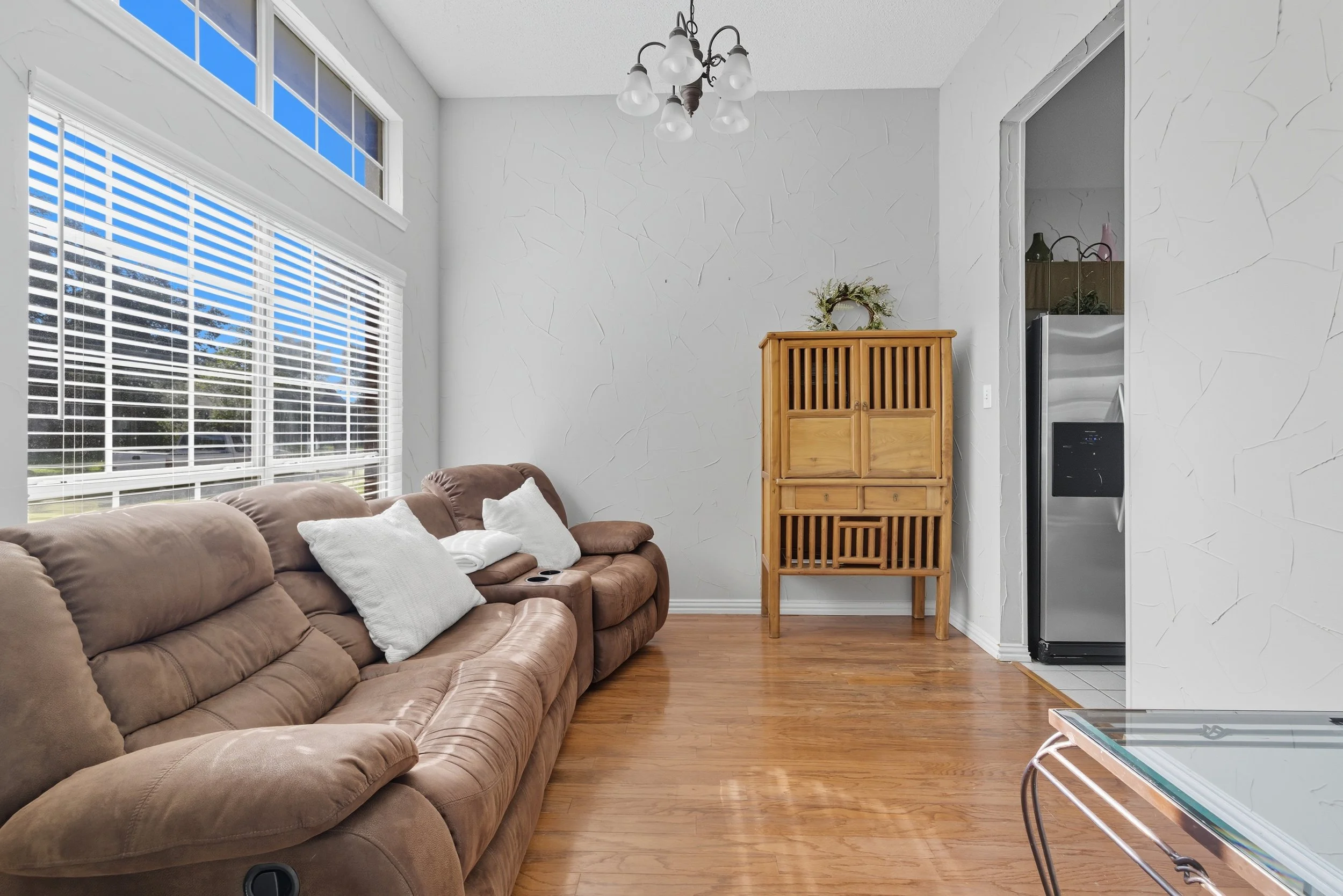 Living room with a brown sectional sofa with pillows, large window with white blinds, wooden flooring, a wooden cabinet, and a partially visible refrigerator in the kitchen area.