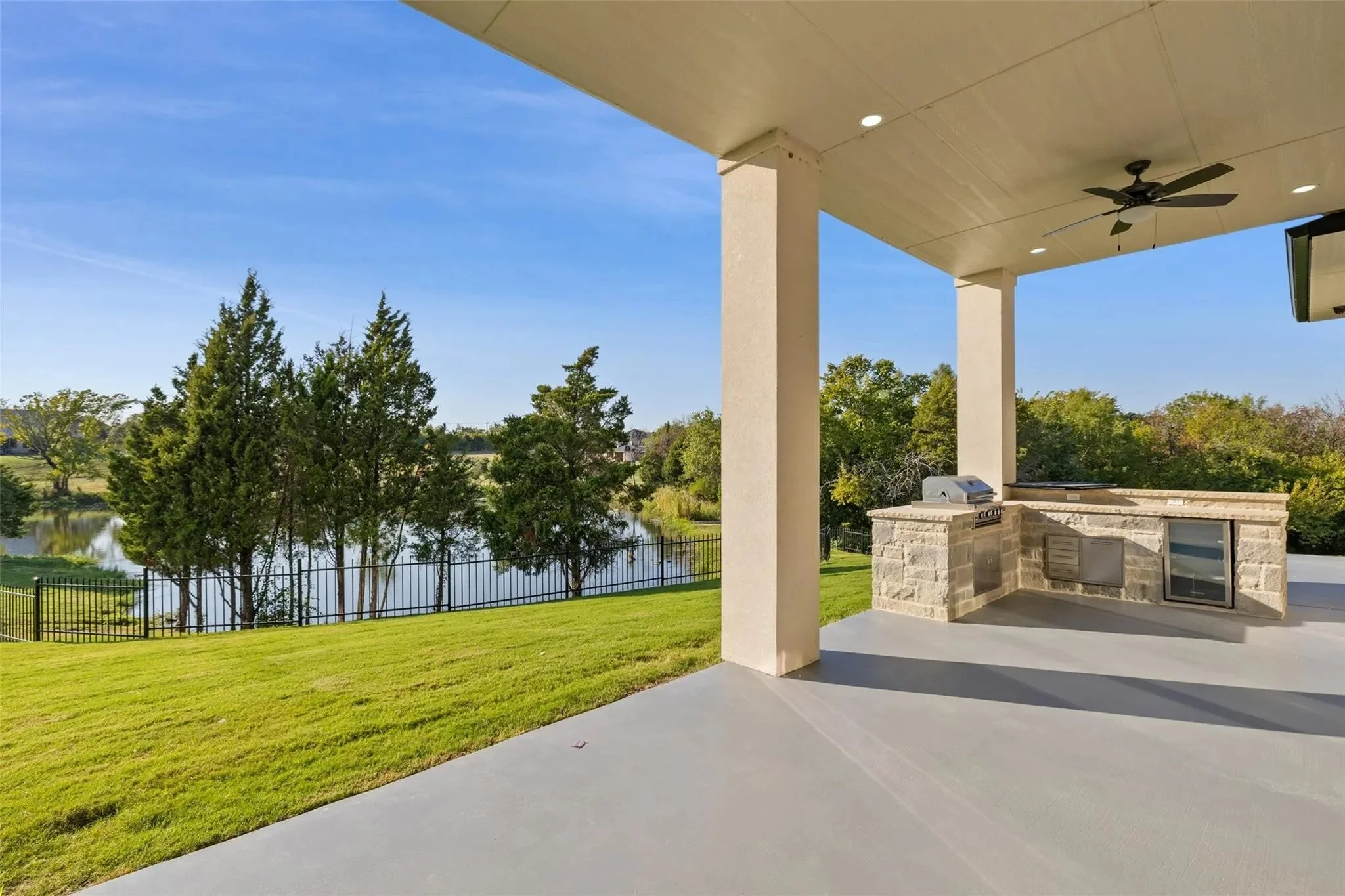 View of a covered patio with a built-in stone outdoor kitchen, ceiling fan, and overlooking a grassy lawn, trees, and a lake, under a blue sky.