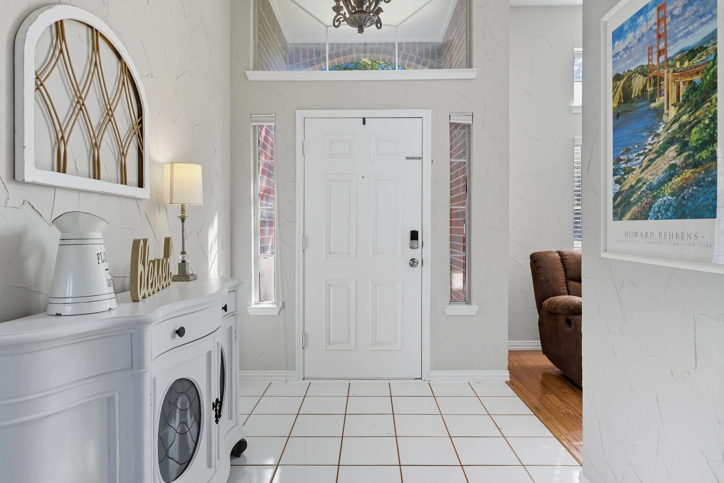Entryway with white door, side windows, white textured walls, tiled floor, and a brown recliner in adjacent room.