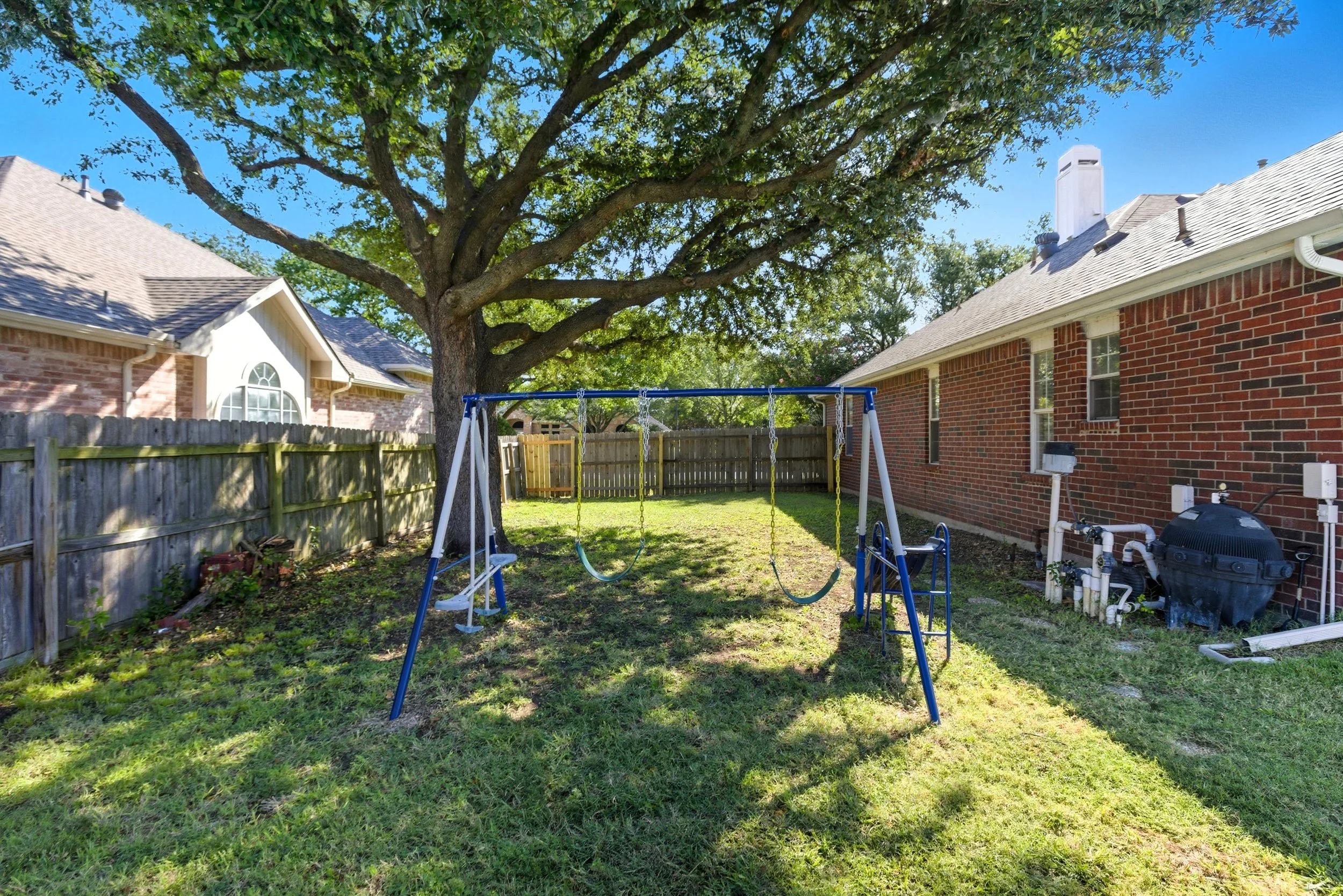 Backyard with a tree, a blue and white swing set with two swings, surrounded by a wooden fence and brick houses.