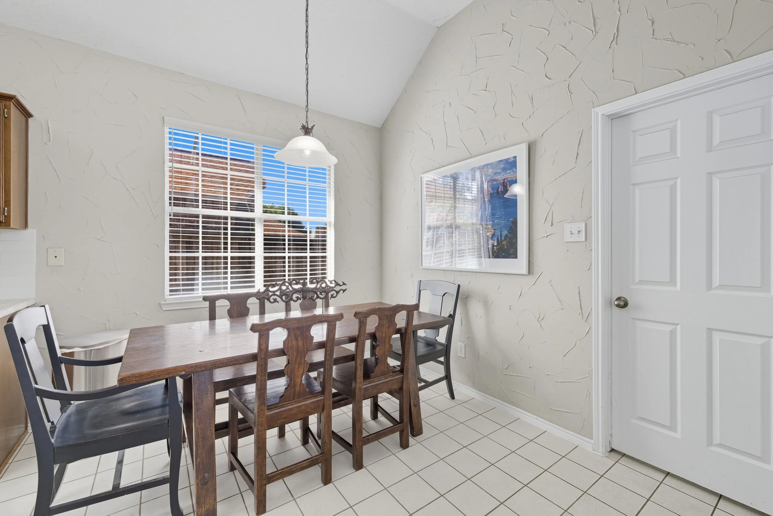 Dining area with a rectangular wooden table, mix of black and wooden chairs, white tile floor, textured beige walls, a window with white blinds, a framed picture on the wall, and a white door.