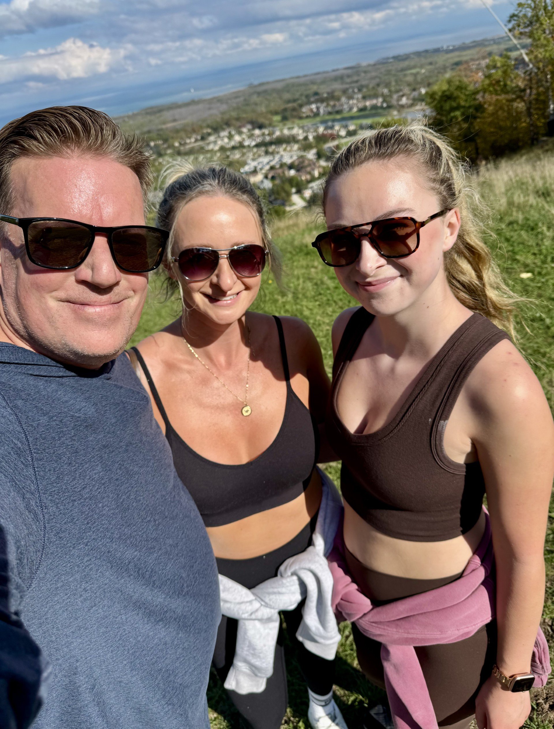 Three people, two women and one man, smiling outdoors with a scenic view of a town, water, and hills in the background, all wearing sunglasses and casual athletic clothing.