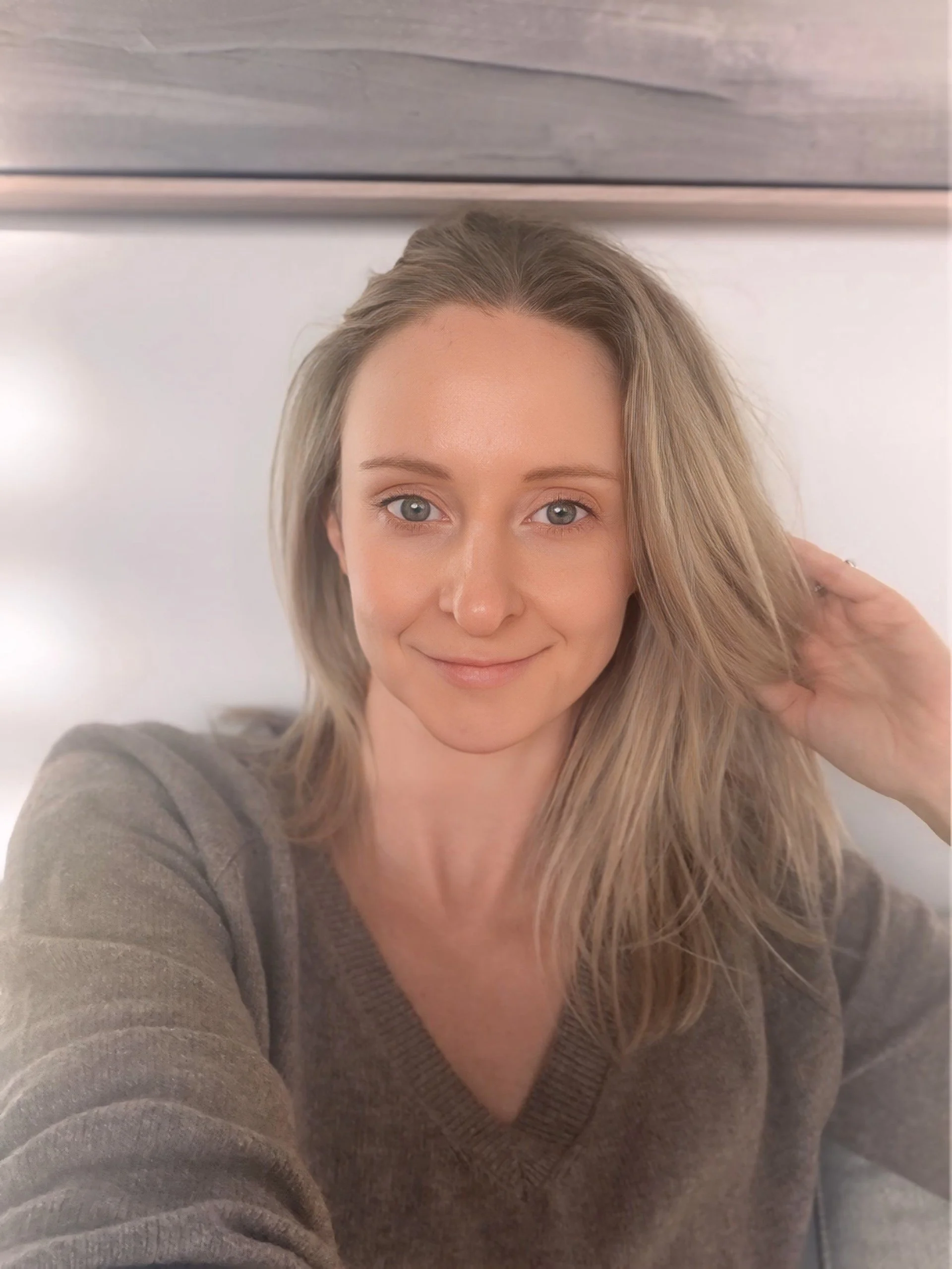 A woman with blonde hair and blue eyes smiling, wearing a brown sweater, touching her hair with her right hand, in a close-up selfie against a light-colored background.