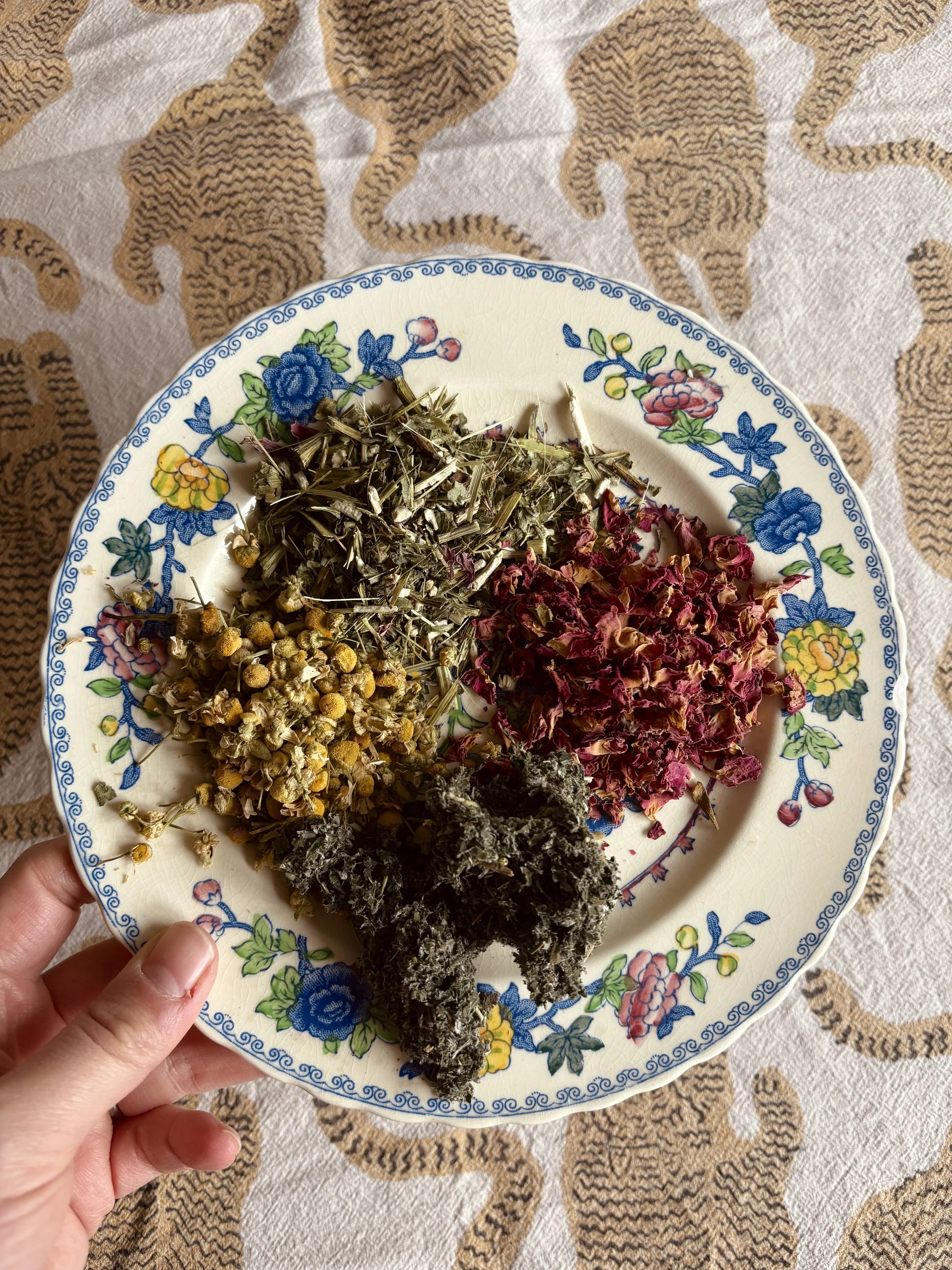 A plate of nourishing and supportive herbs, including chamomile, raspberry leaf and rose, on a colourful patterned plate.